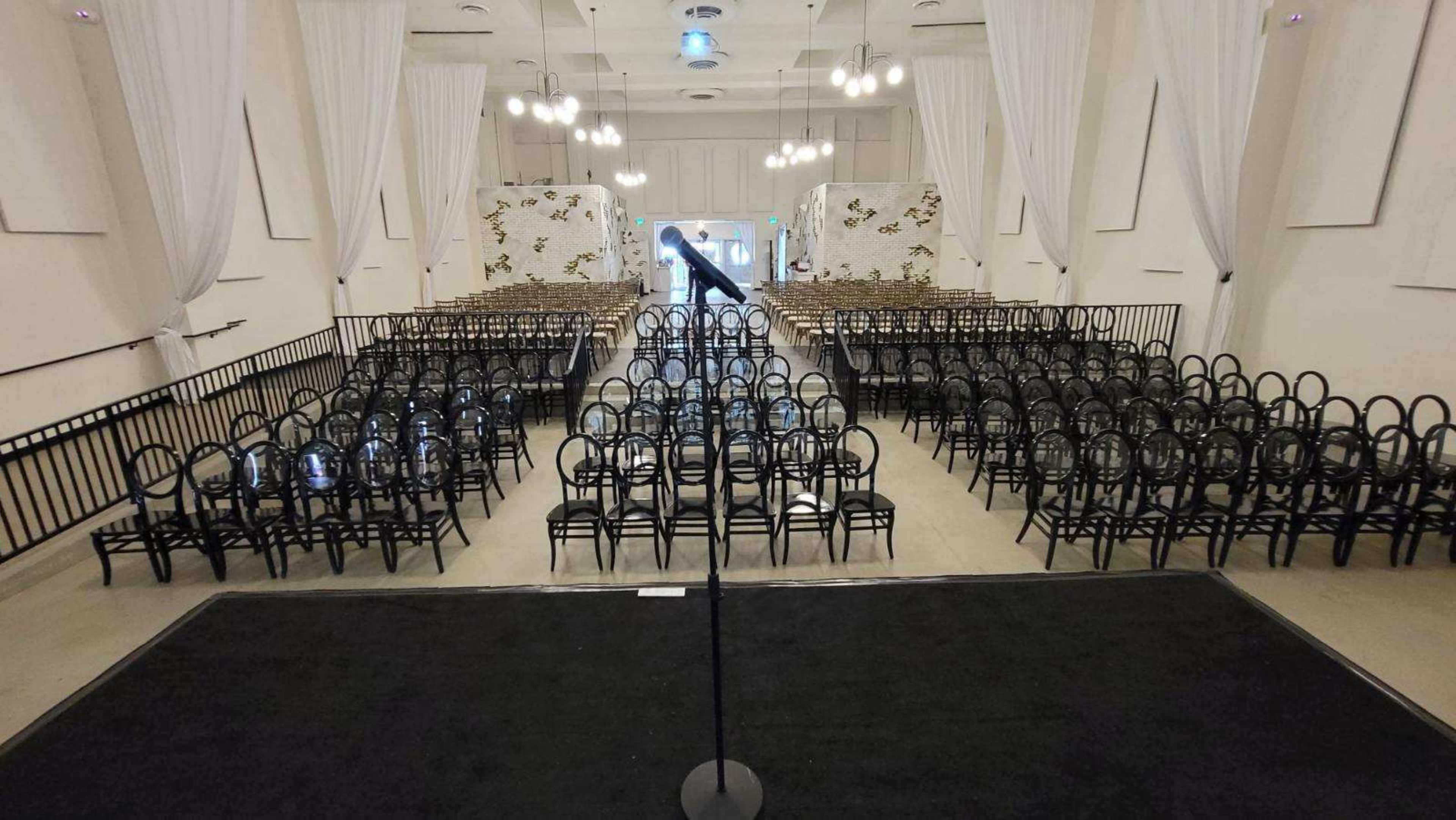 An empty auditorium features rows of black chairs arranged facing a stage with a microphone stand at the front.