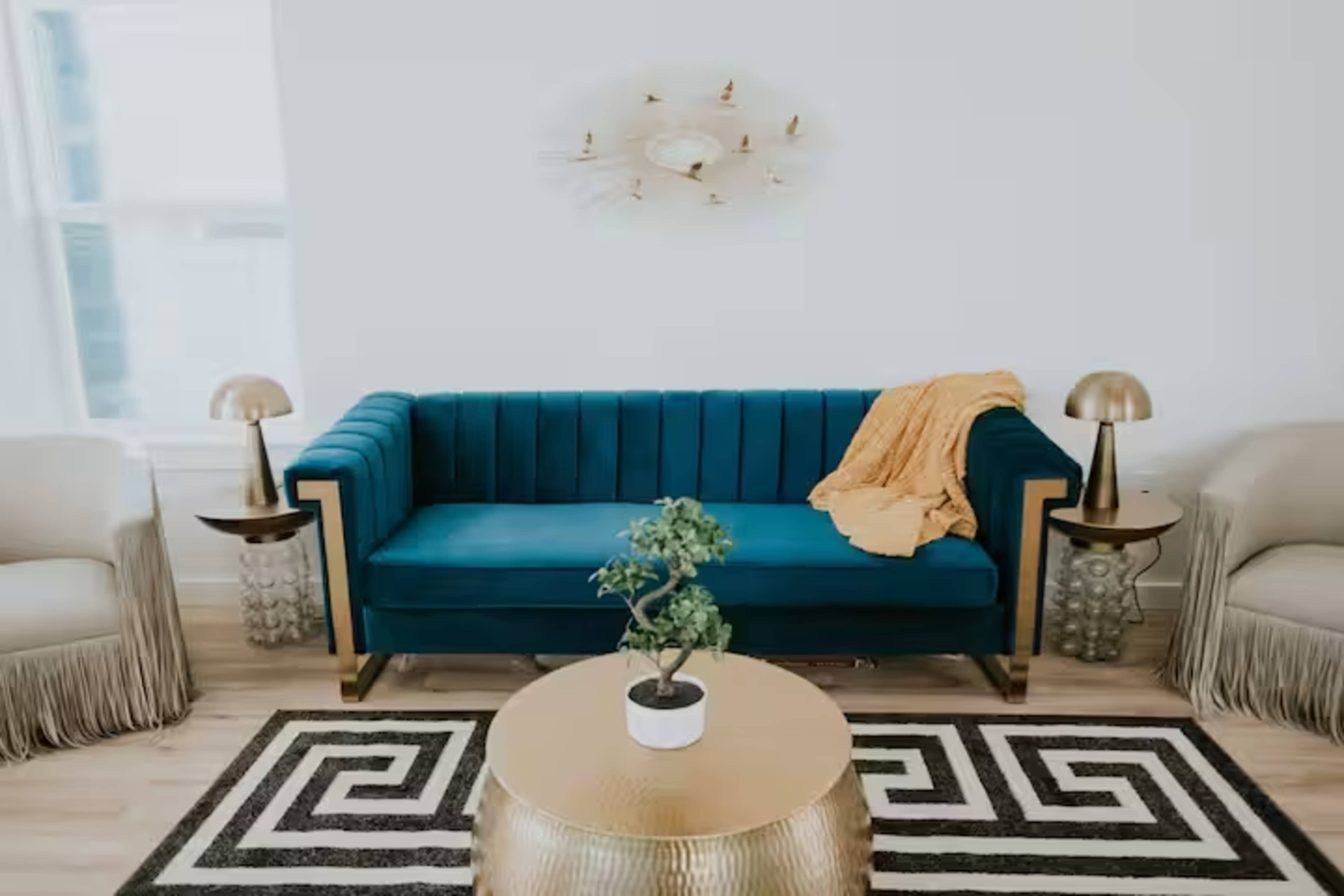 The scene features a modern living room with a teal sofa, two armchairs, a round gold coffee table, and decorative lamps on either side of the sofa.