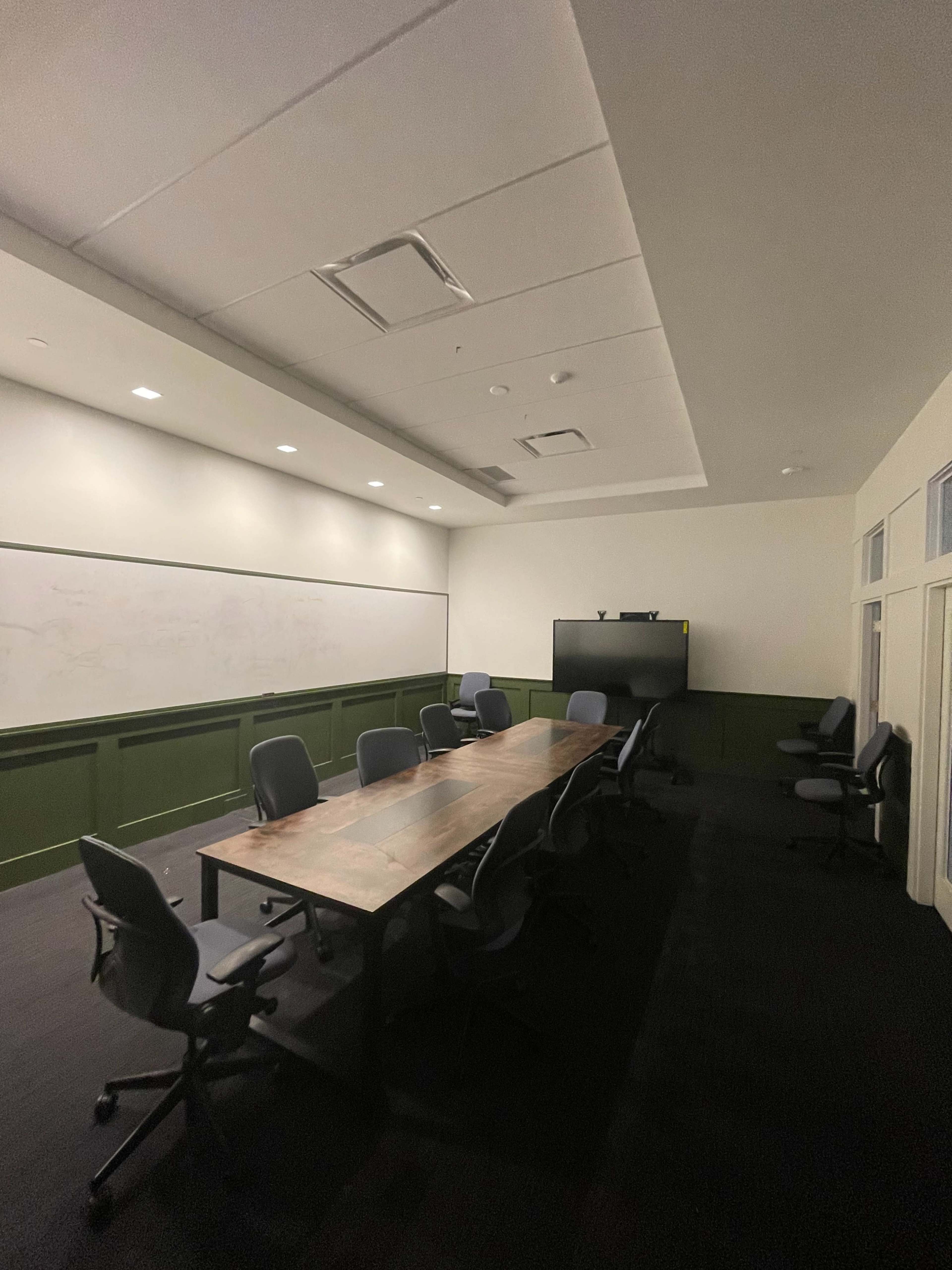 A conference room features a long wooden table surrounded by chairs, with a blank whiteboard and a television screen on the wall.