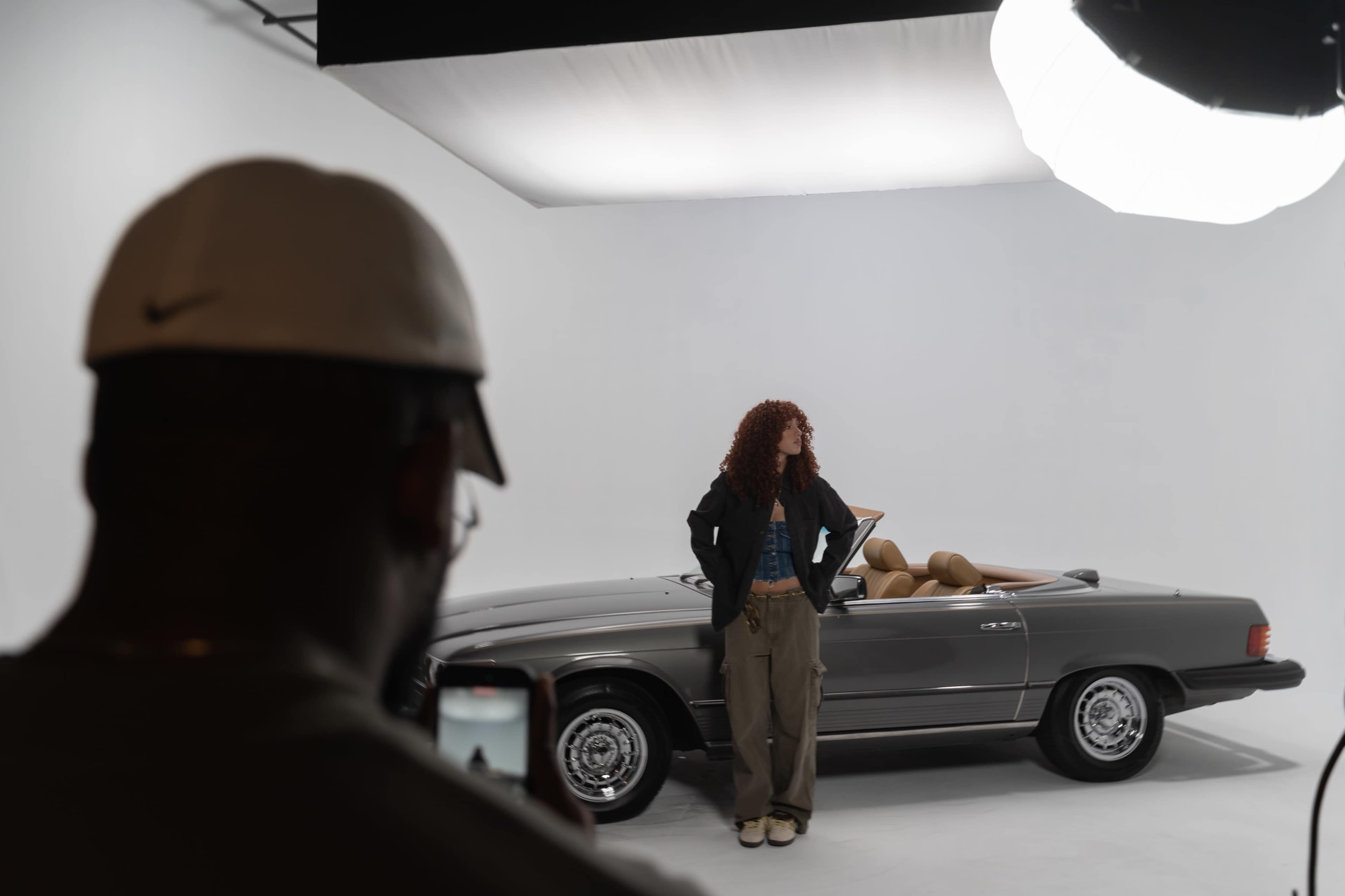 A woman poses next to a convertible car in a photo studio while a person takes a picture of her.