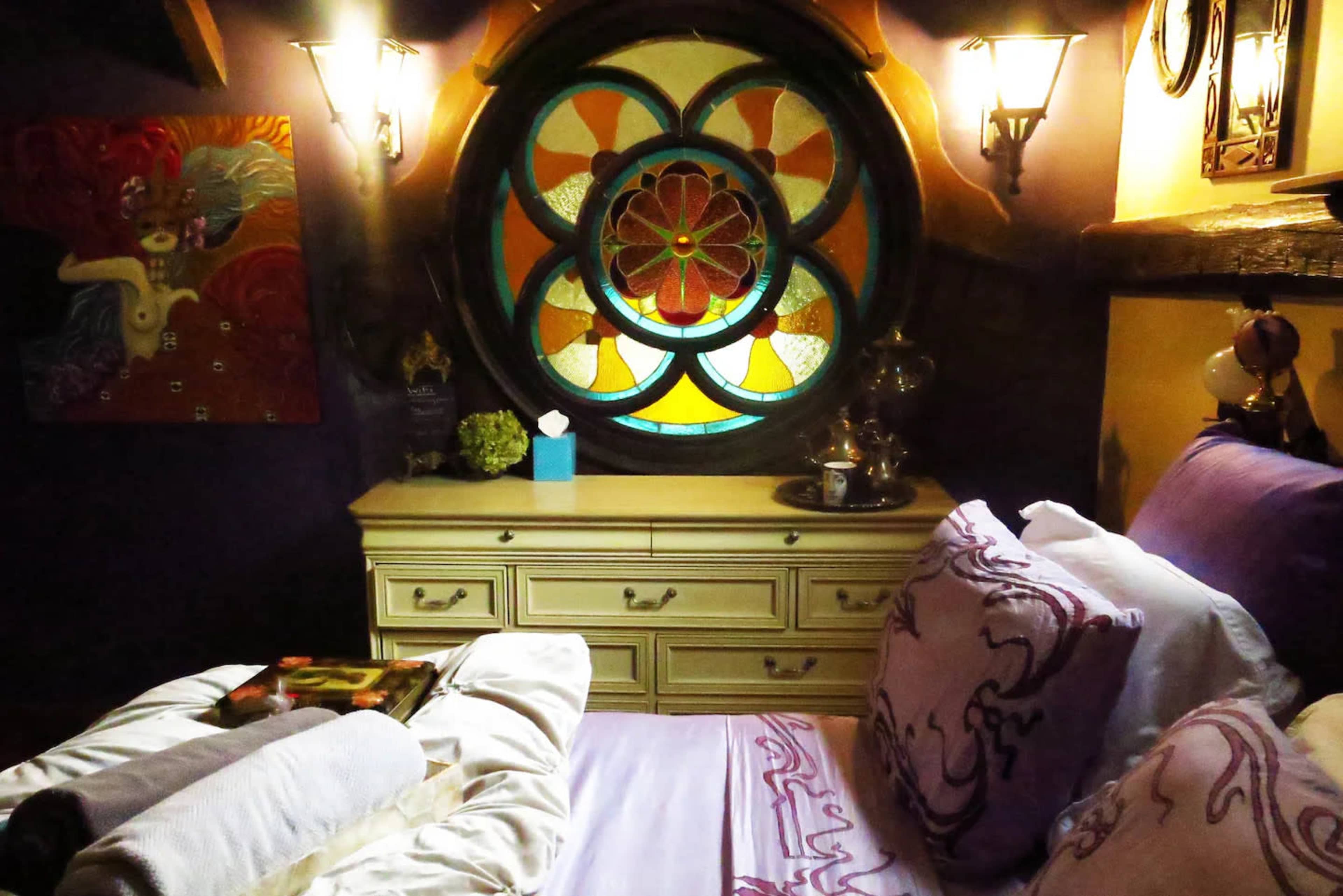 The image shows a cozy bedroom with a large, colorful stained glass window and decorative lighting.