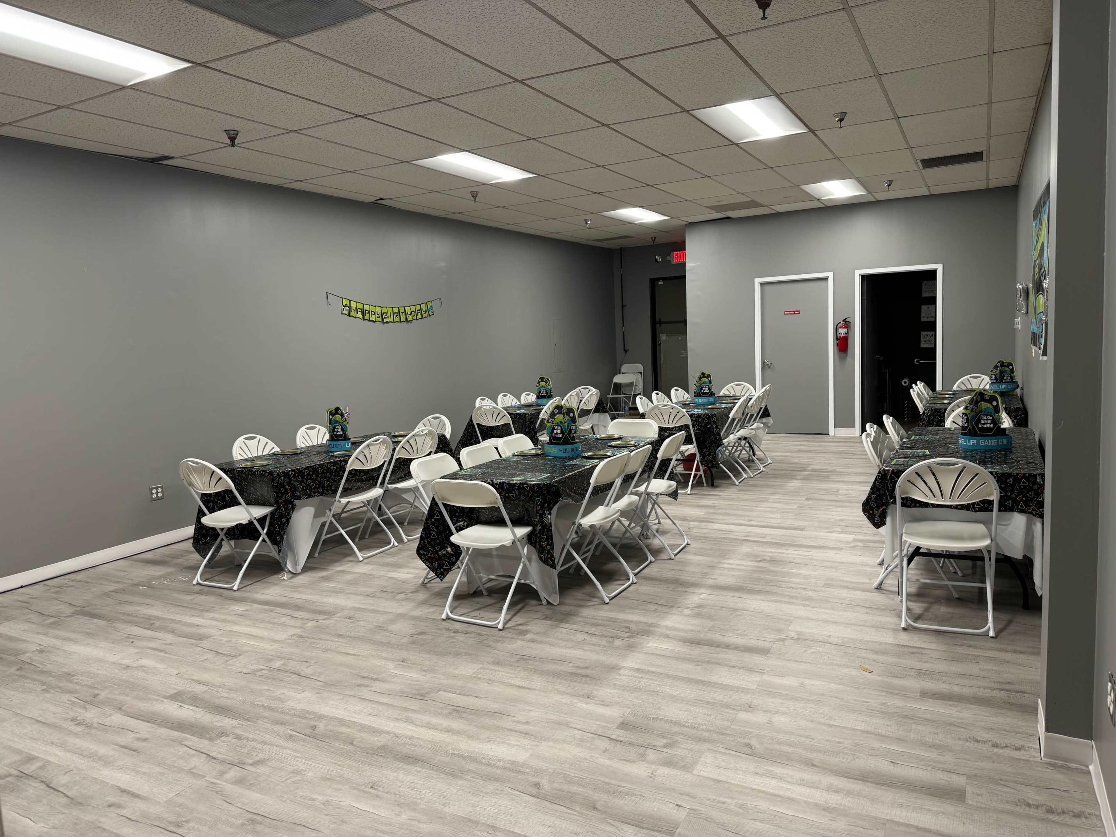 A well-lit event space featuring several round tables covered with black tablecloths and arranged neatly with chairs around them.