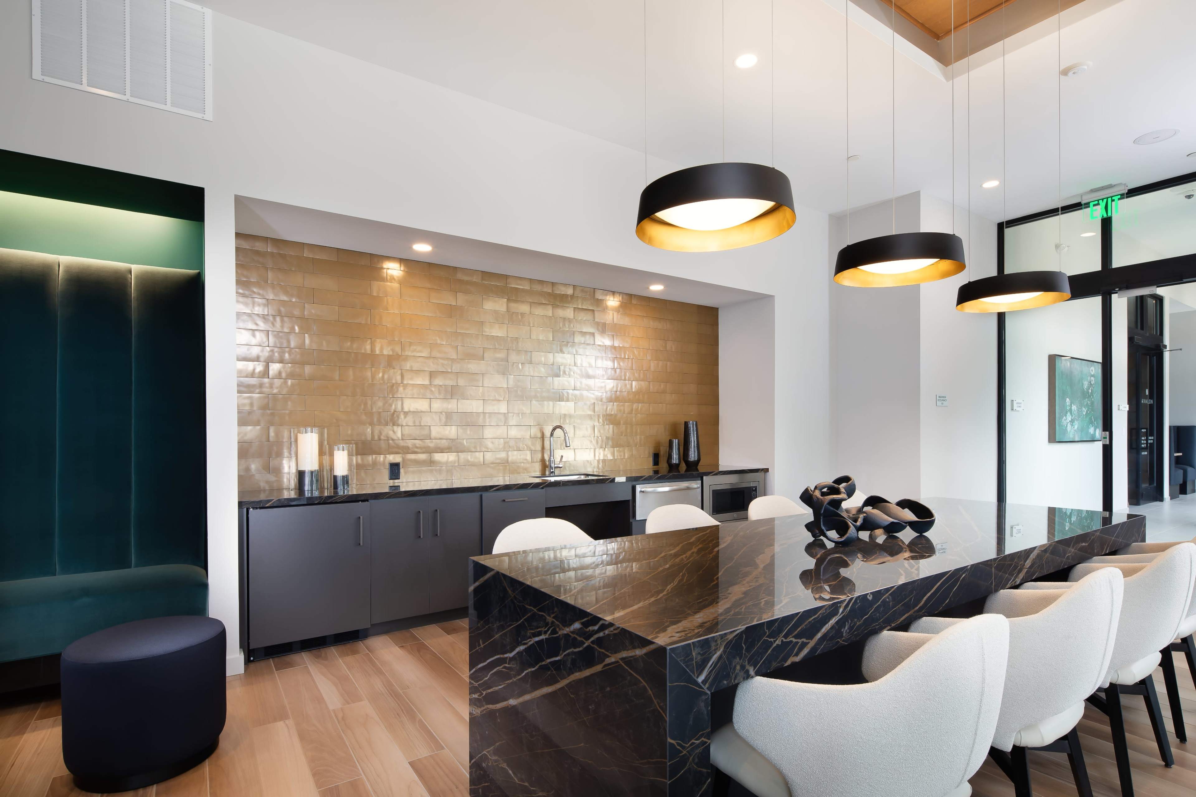 A modern kitchen area with a large black marble island, light-colored upholstered chairs, and gold backsplash tiles, illuminated by three pendant lights.