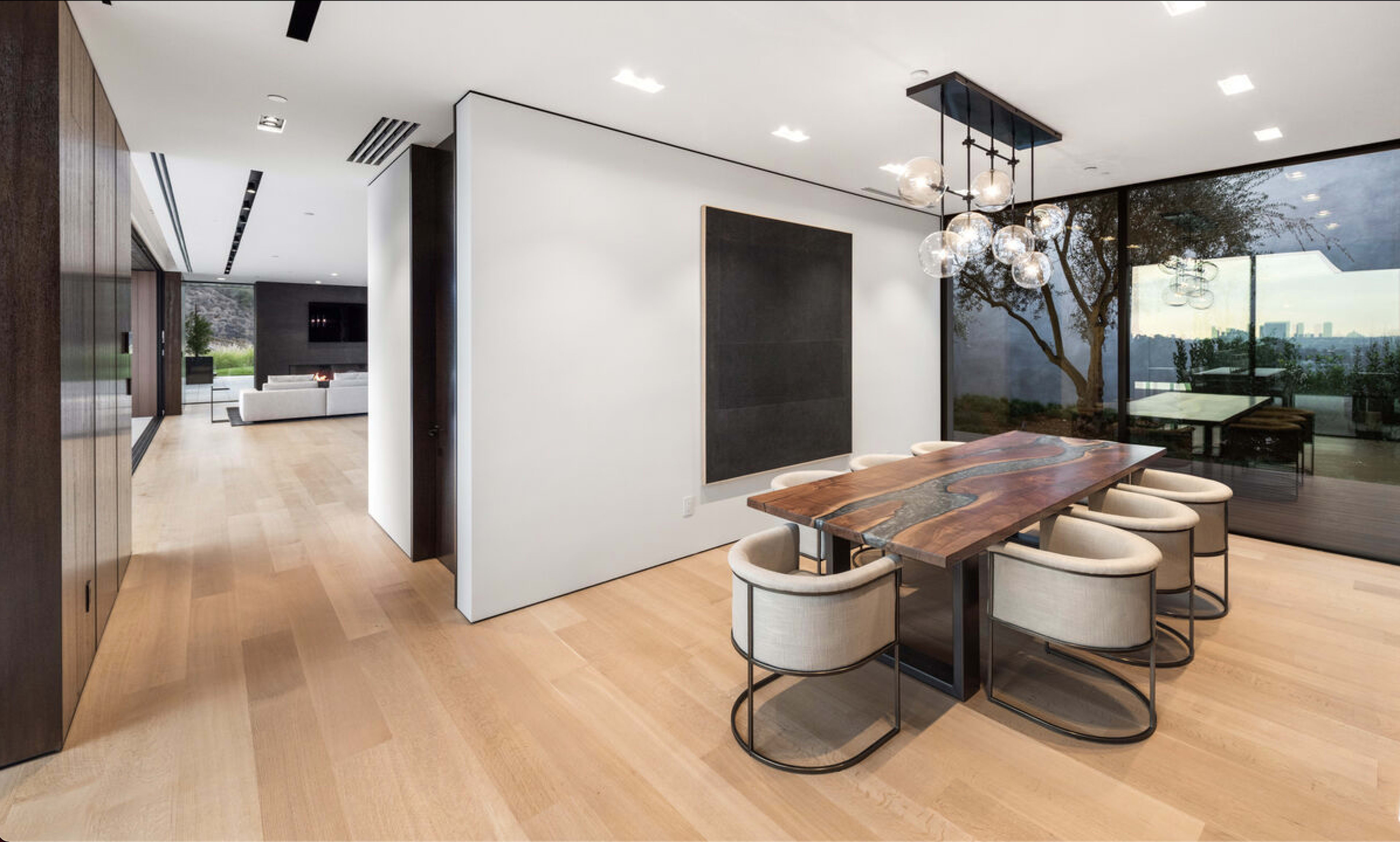A modern dining area with a wooden table surrounded by circular upholstered chairs, situated next to large glass windows that provide a view of the outdoors.