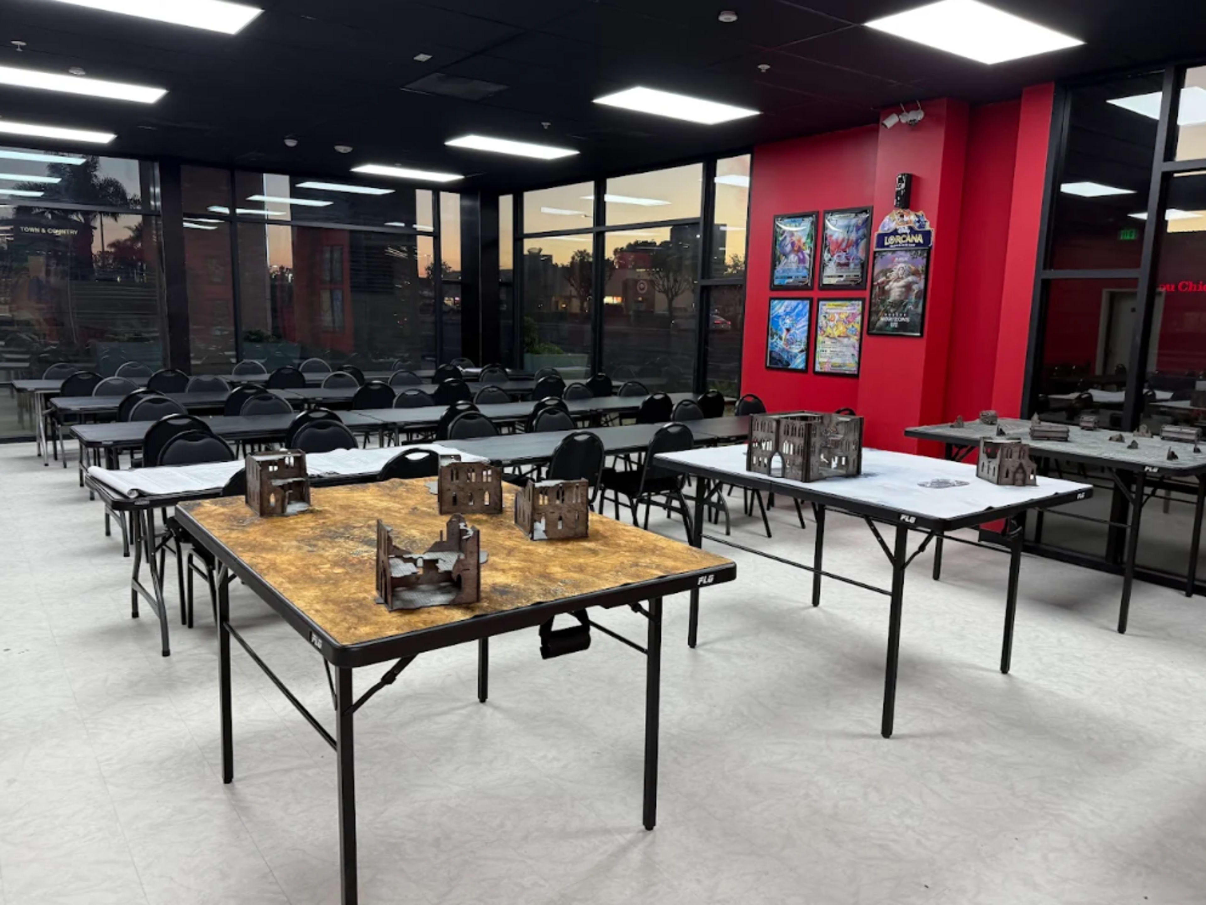The image shows a spacious room with multiple tables arranged for a gaming event, featuring small model buildings on some tables and an array of empty chairs.