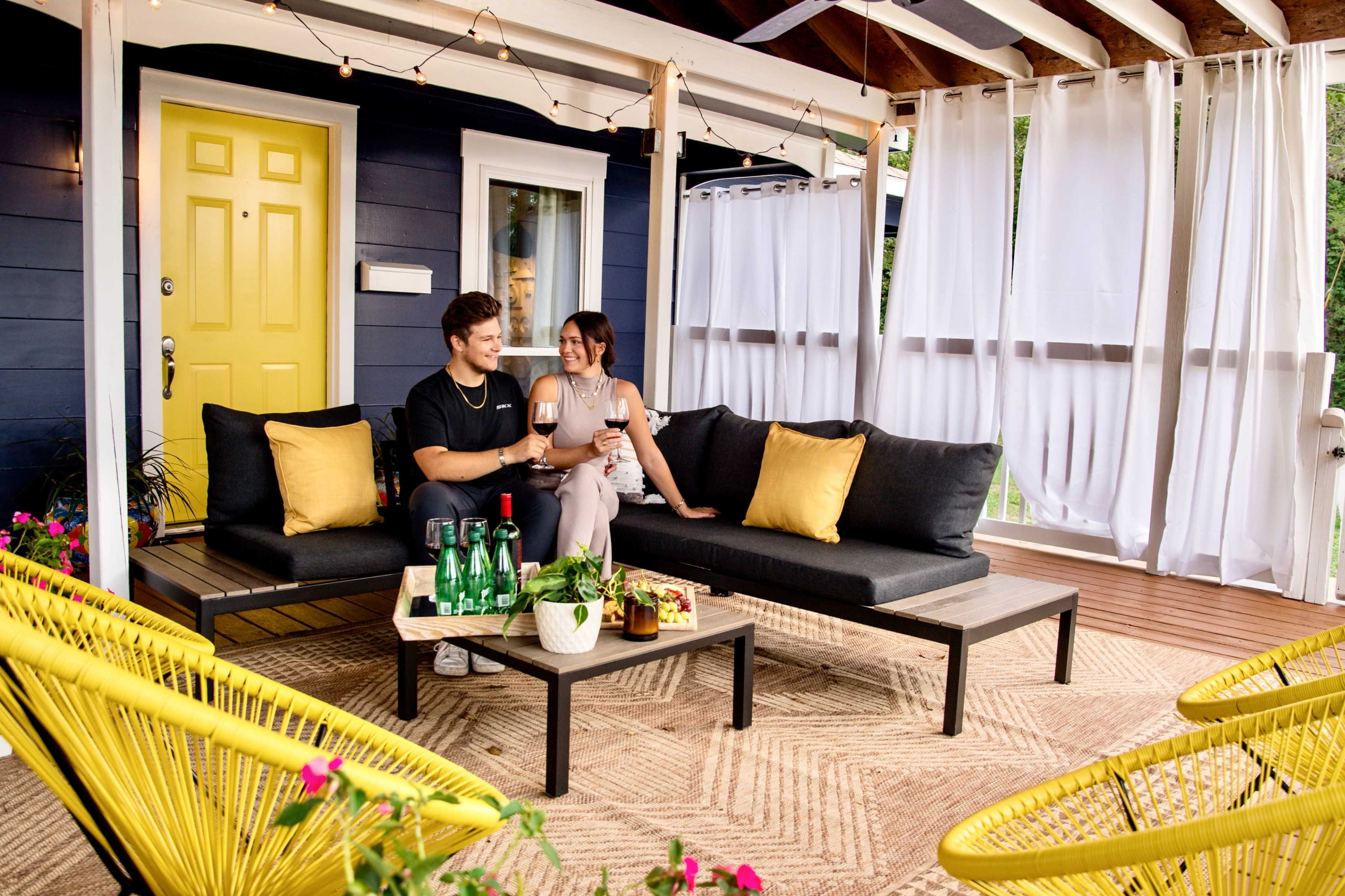 A couple sits on a patio with black couches and yellow pillows, enjoying drinks at a table on a deck decorated with plants and white curtains.