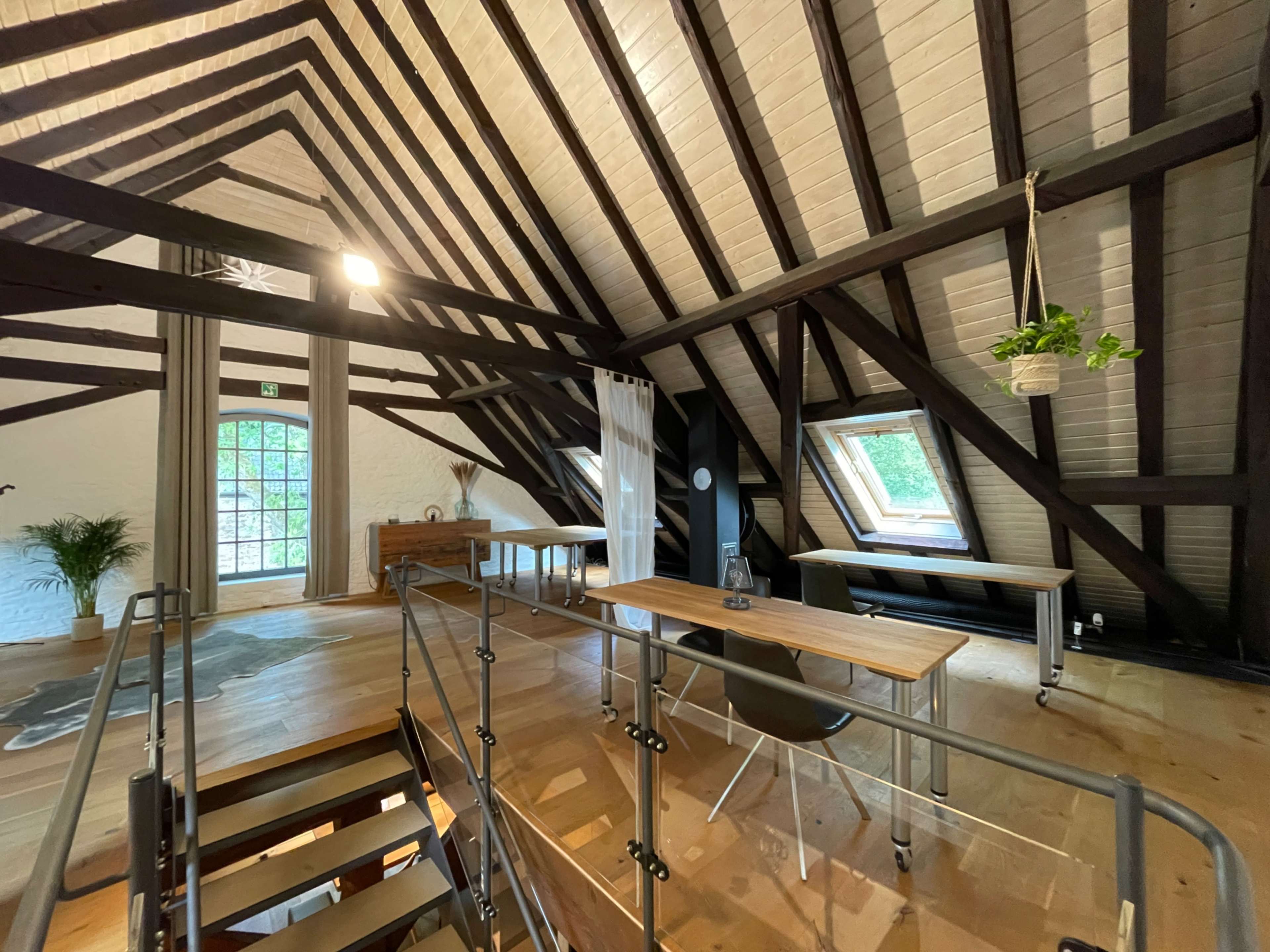 The image shows a spacious, open loft with wooden beams and large windows, featuring a minimalist workspace and a staircase.