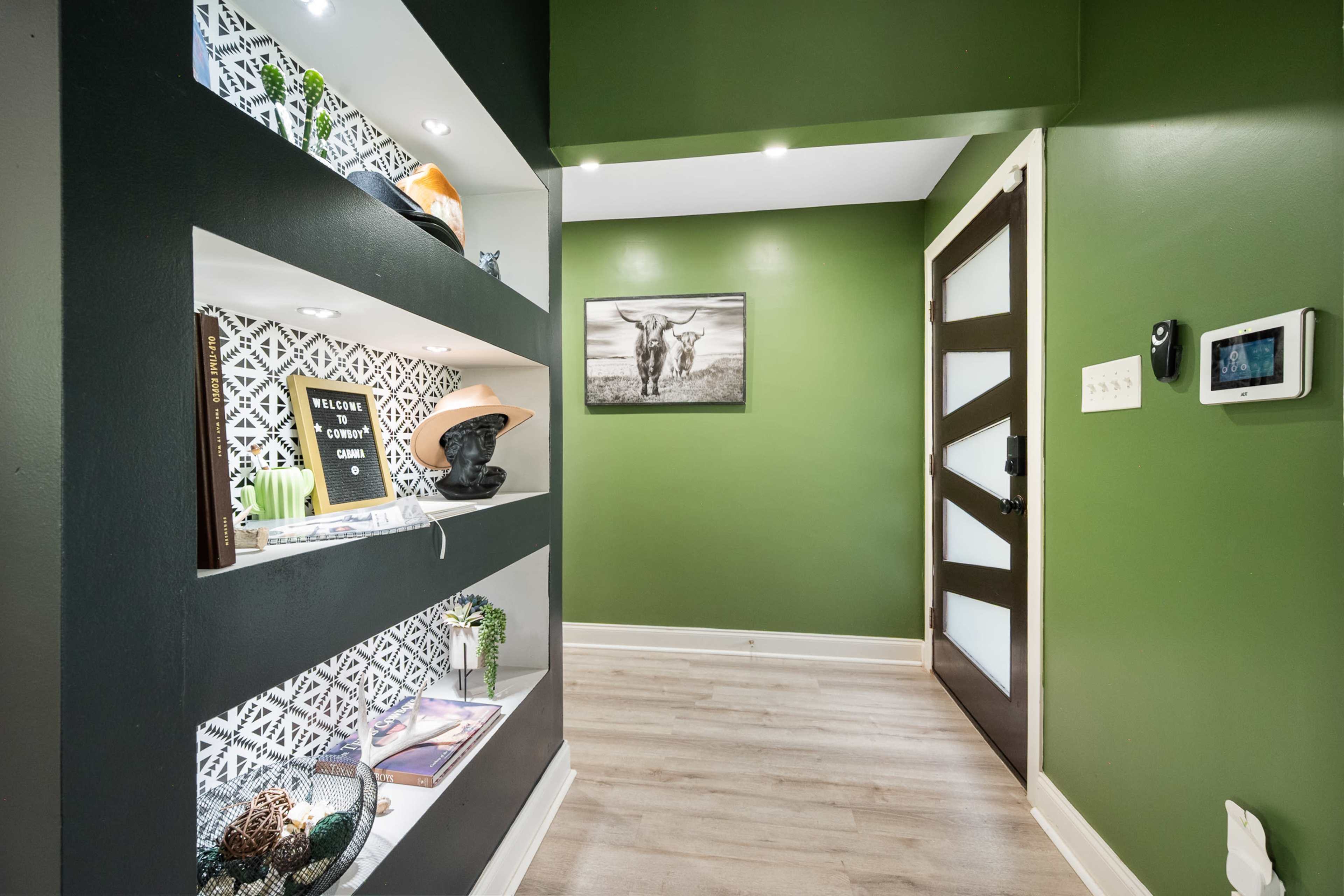 The image shows a hallway with dark green walls, a shelf displaying decorative items, and a door featuring a contemporary design.