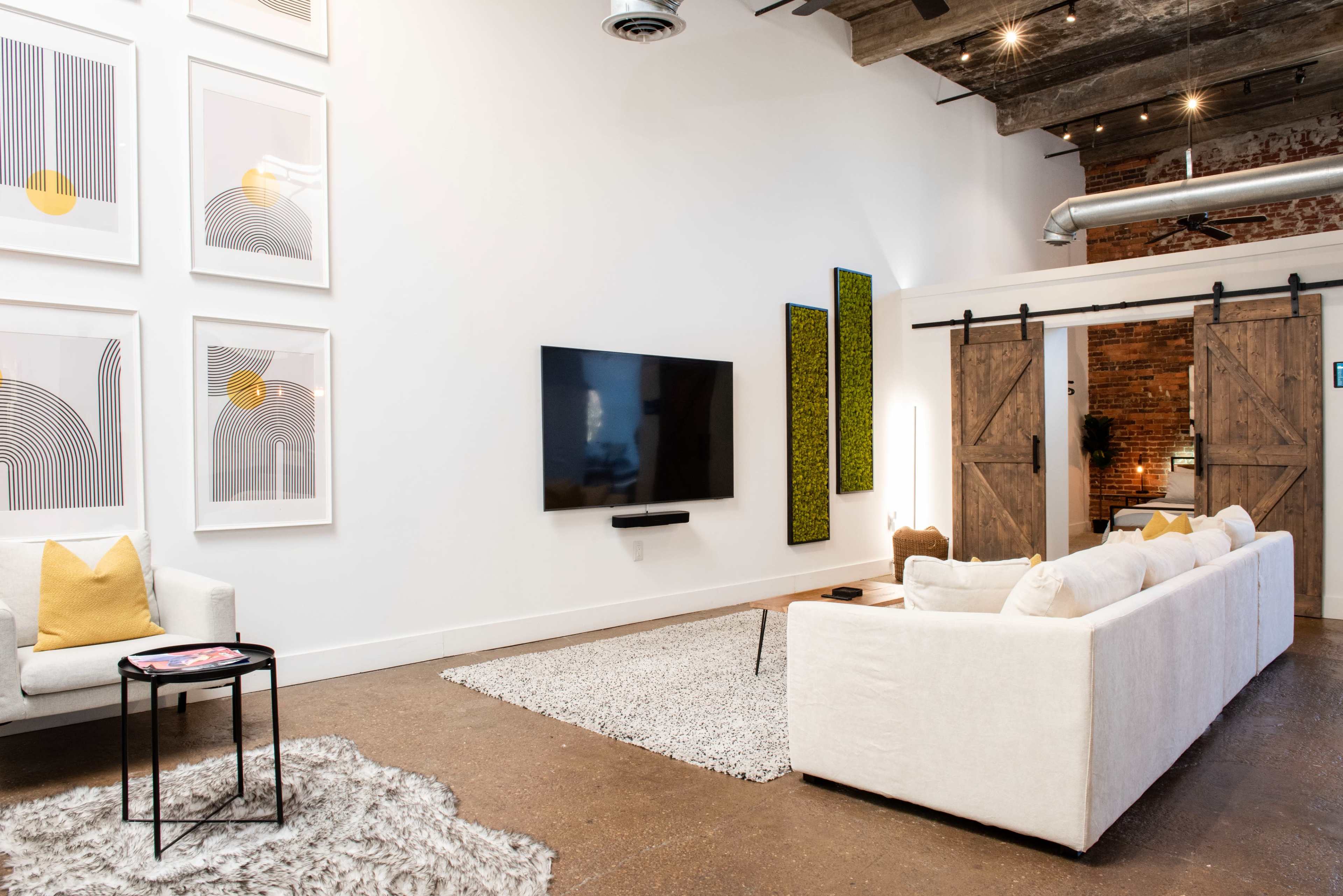 A modern living room with a large white sectional sofa, a black coffee table, a flat-screen TV mounted on the wall, and abstract art framed on the wall, complemented by sliding barn-style wooden doors.