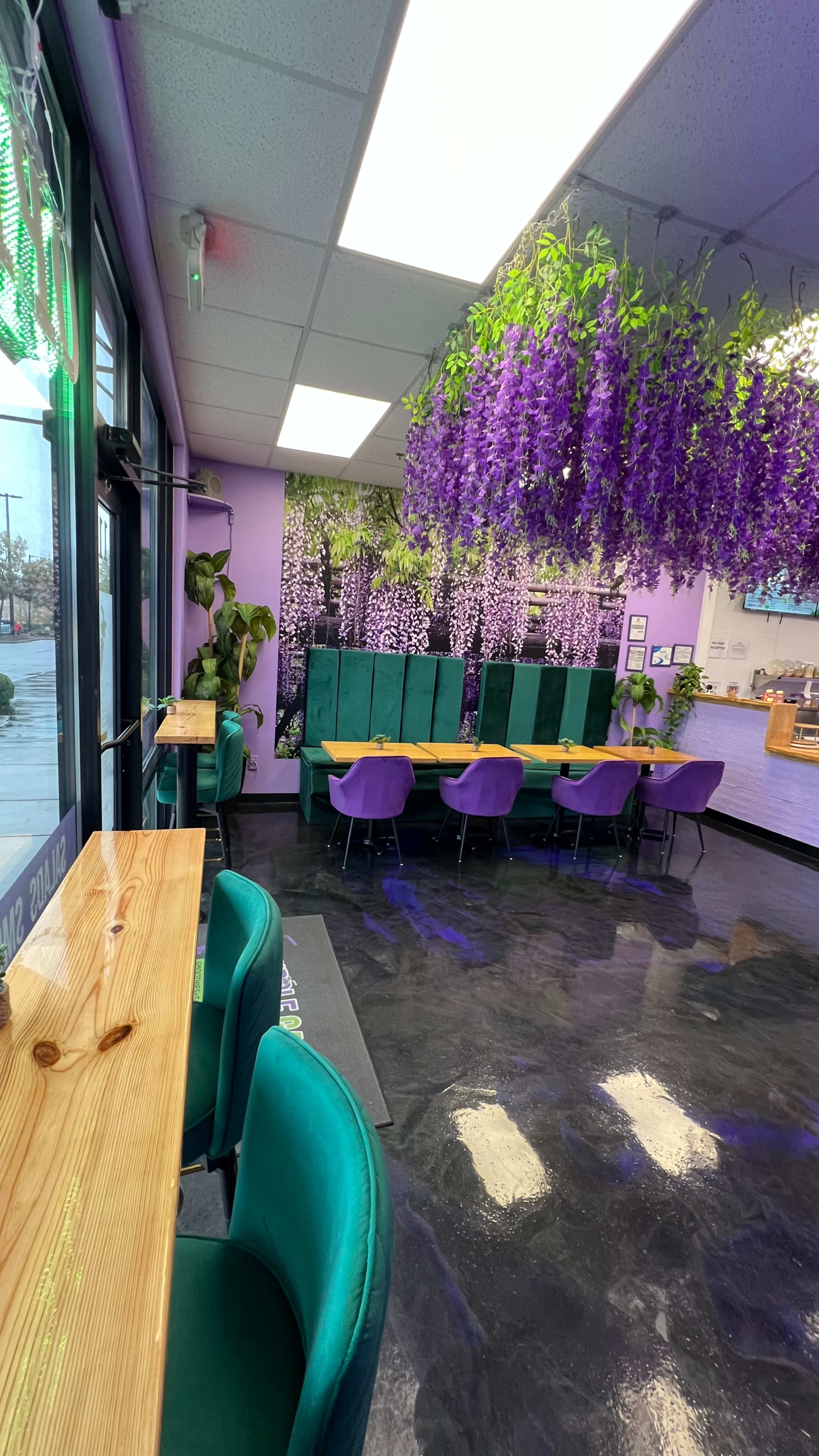 The image shows a brightly colored café interior with green accent chairs, purple tables, and purple hanging plants, creating a lively atmosphere.