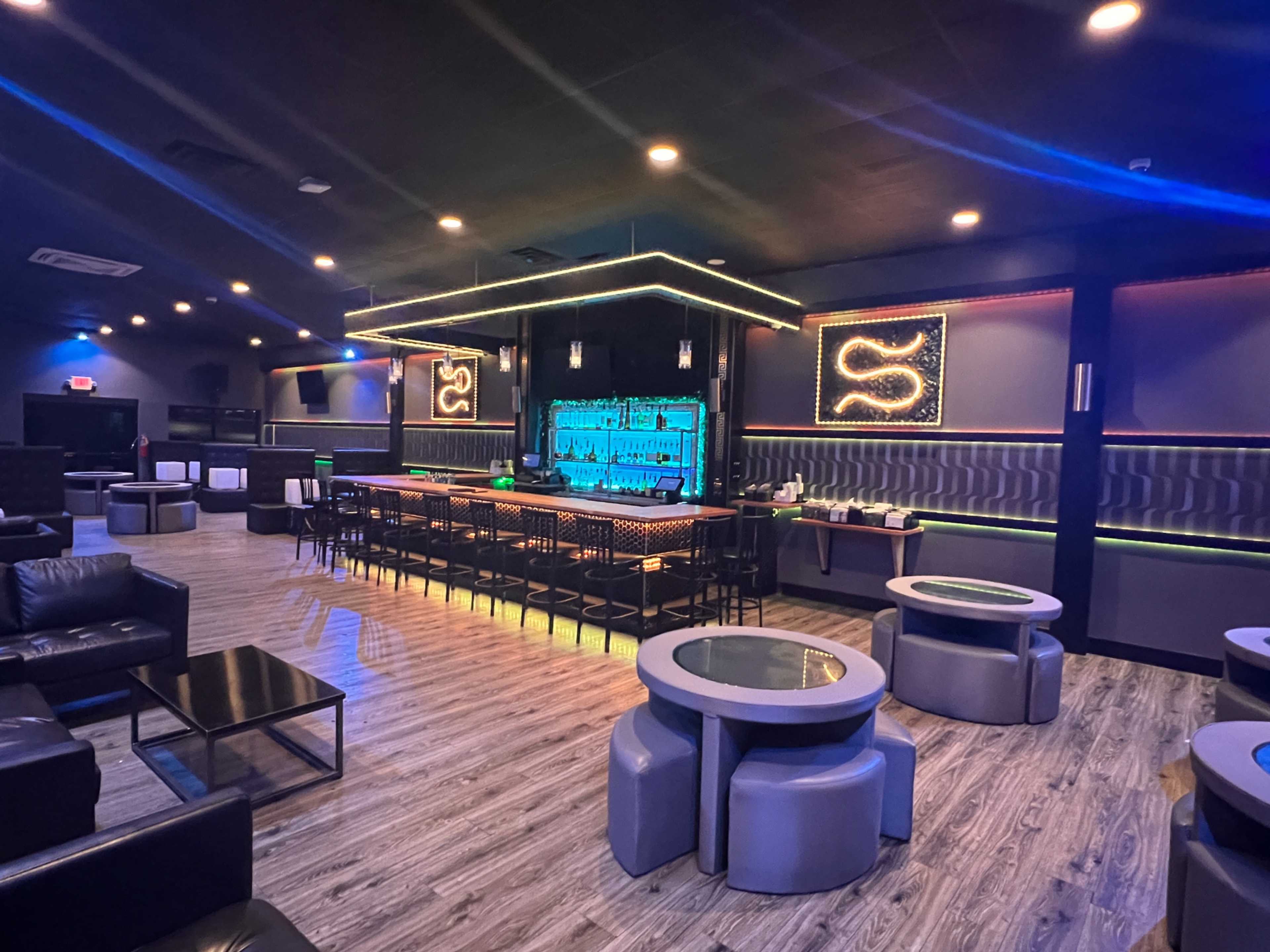 A modern lounge interior with a sleek bar, illuminated by blue lighting, surrounded by black leather seating and circular tables.