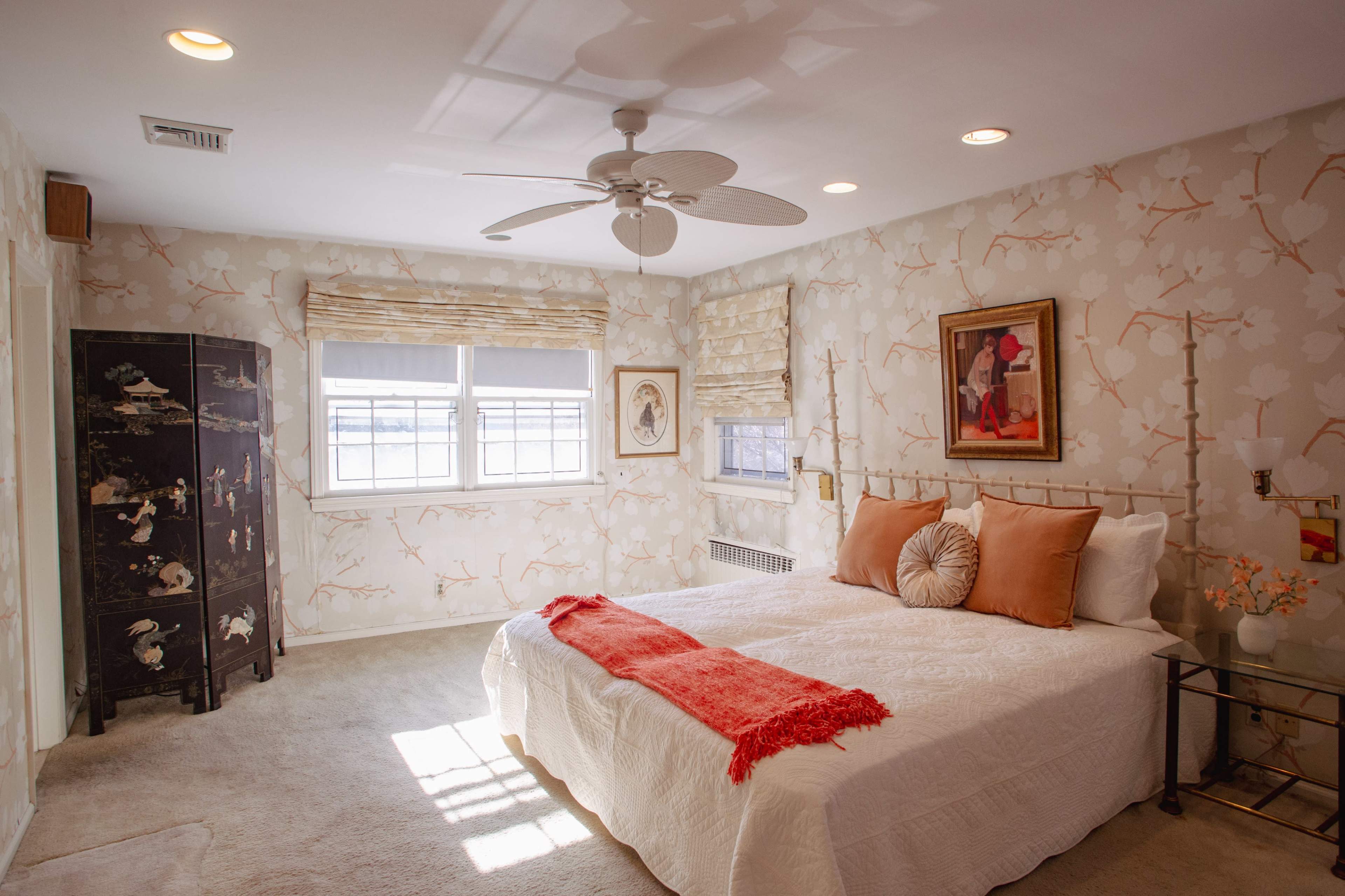 The bedroom features a white bed with orange accent pillows, light-colored walls adorned with floral wallpaper, and a decorative black cabinet in the corner.
