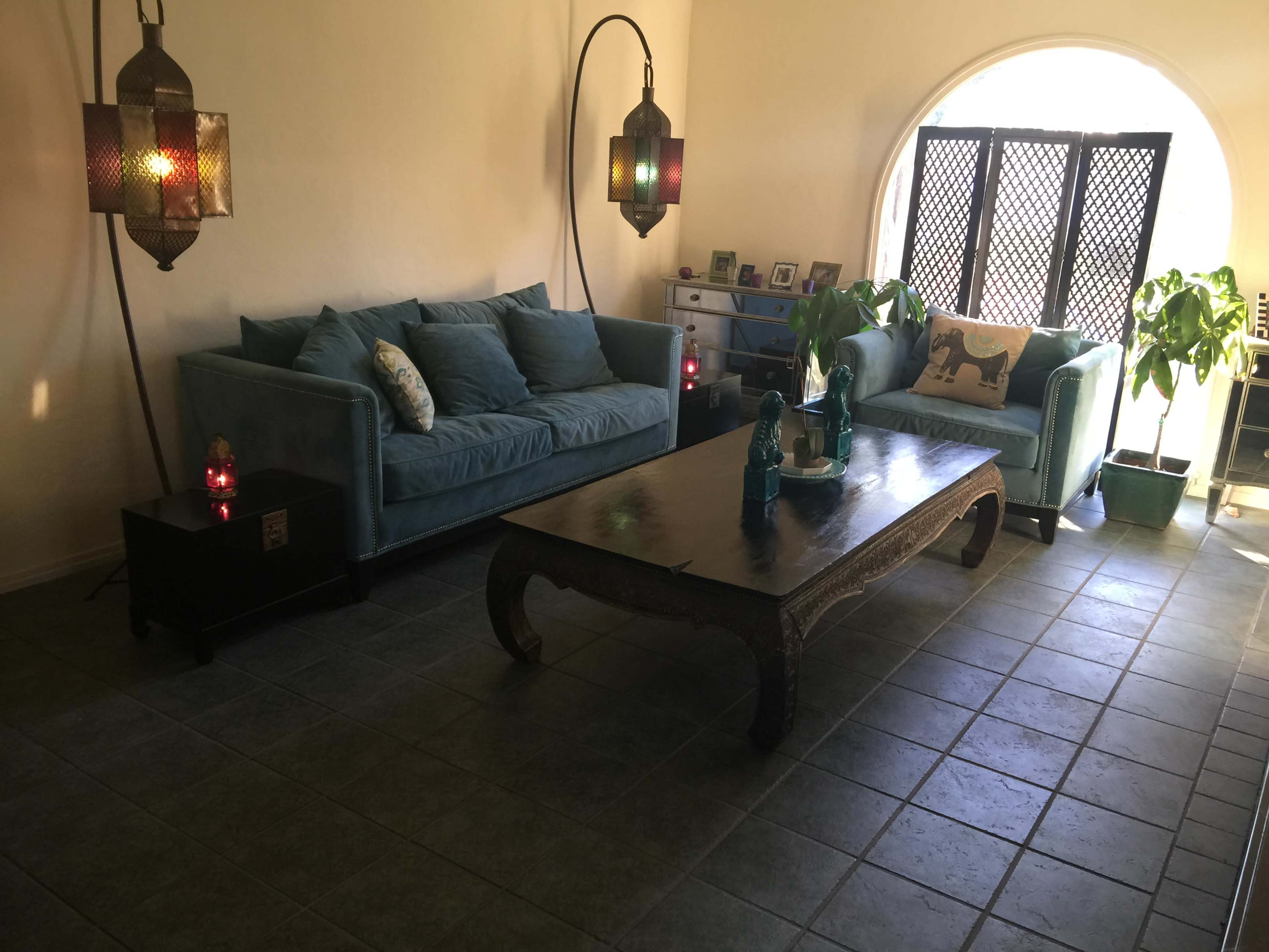 The image shows a living room with two blue sofas, a wooden coffee table, and decorative lamps, featuring a large round window and potted plants.