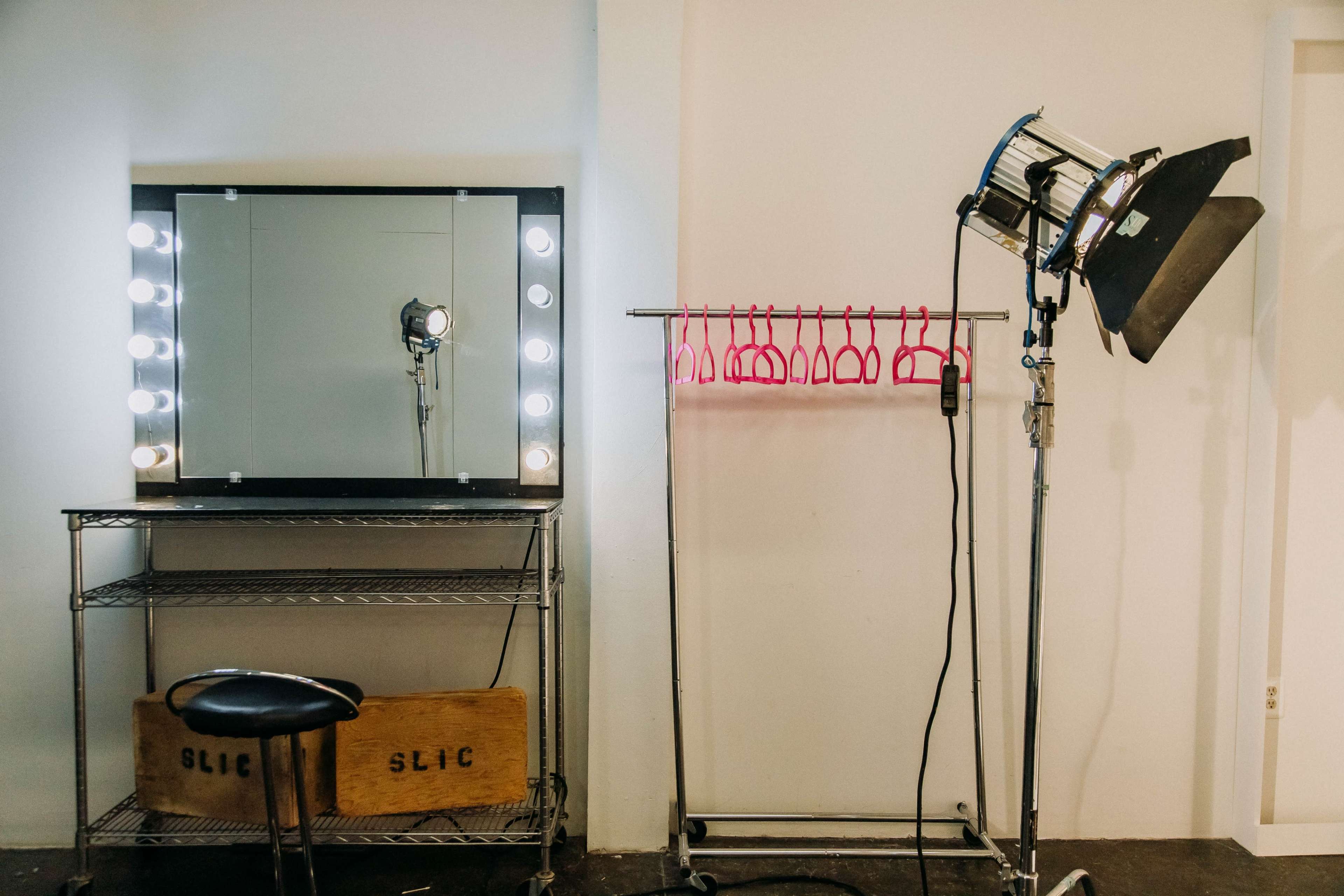 A makeup station with a mirror and bright lights, next to a clothing rack with pink hangers and a lighting setup on a stand.