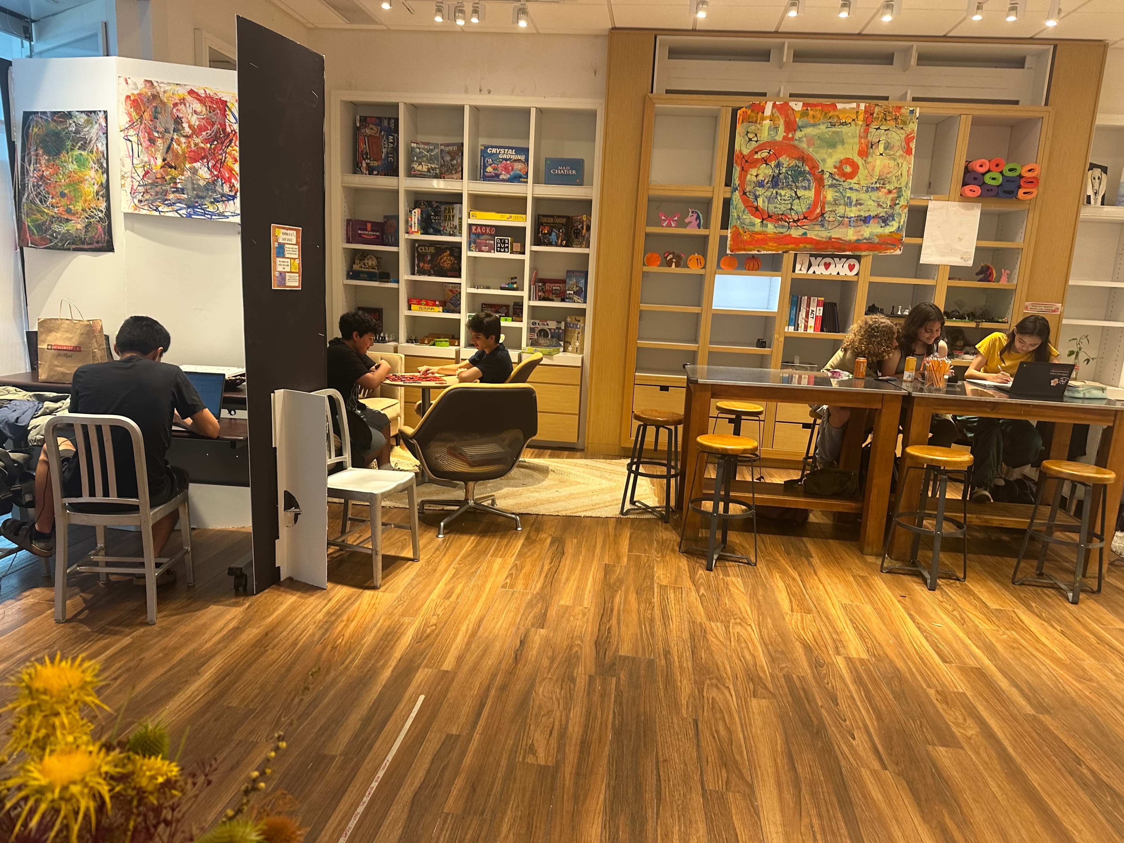 The image shows a brightly lit co-working space with people engaged in various activities at tables and a lounge area, surrounded by colorful artwork and shelves.