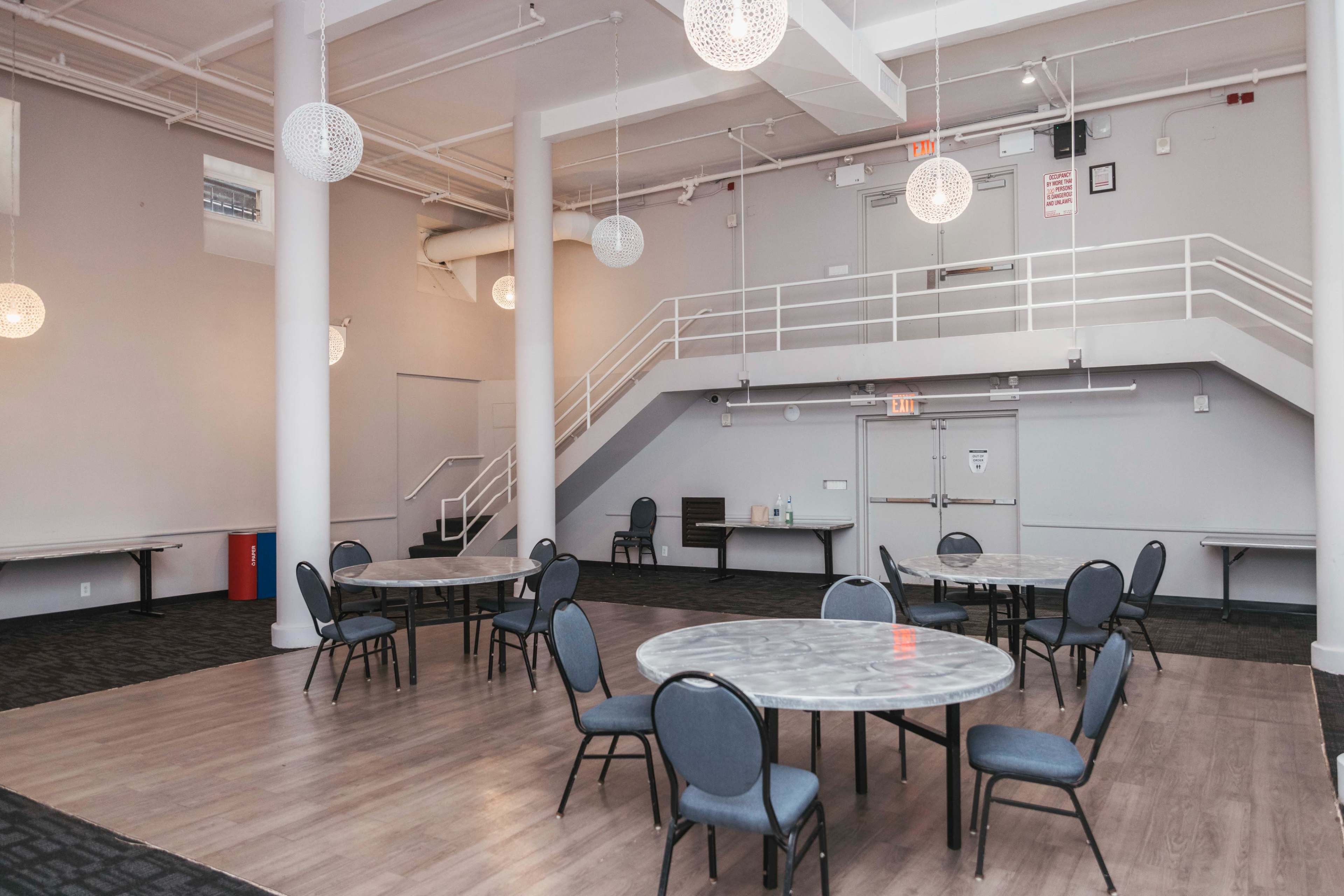 The image shows a spacious indoor area featuring multiple round tables with chairs, illuminated by decorative hanging light fixtures, and a staircase leading to an upper level.