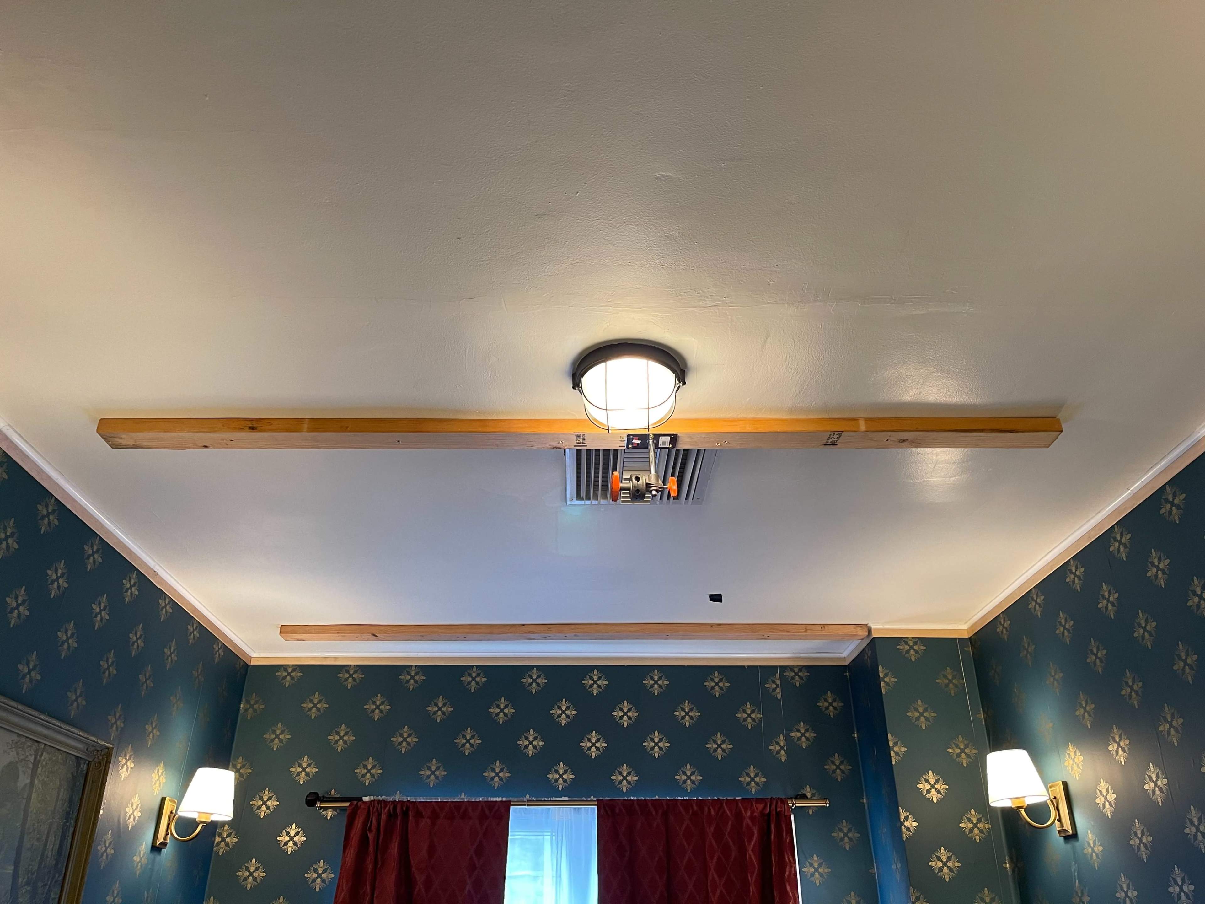The image shows a room with a blue patterned wallpaper, featuring wooden beams on the ceiling and a central light fixture, flanked by two wall sconces.