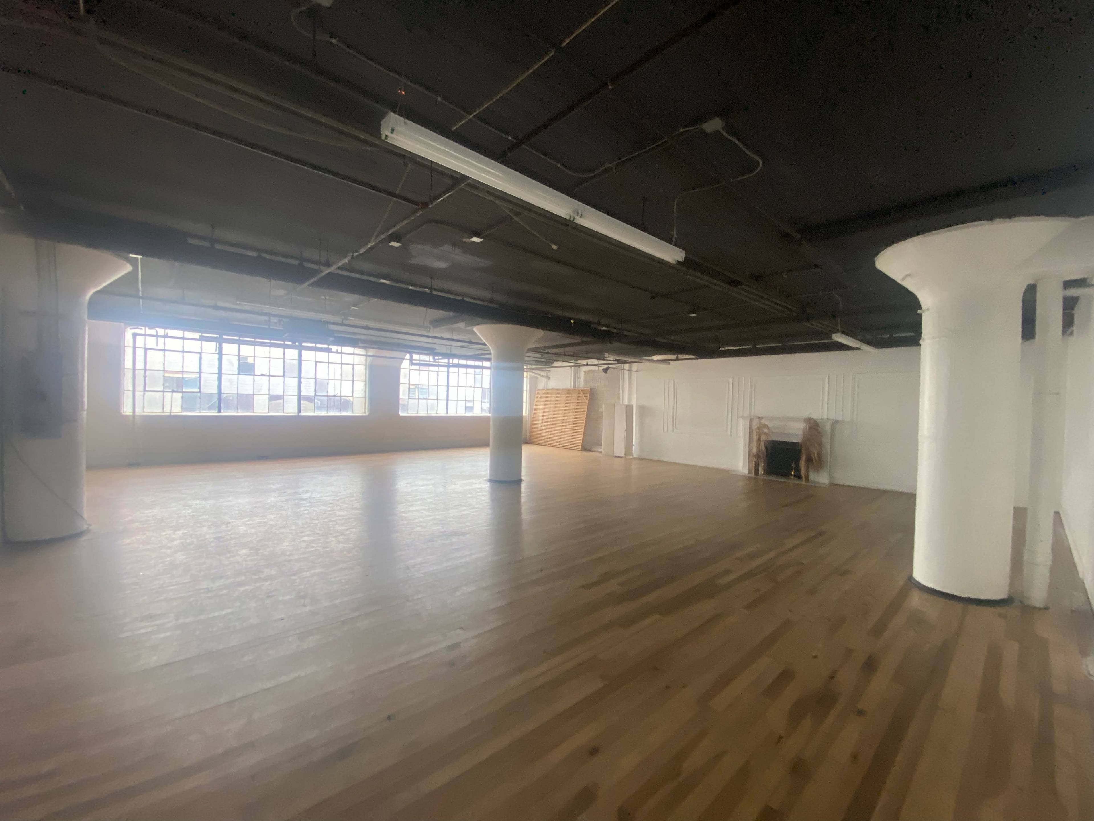 An empty, spacious room with hardwood flooring, large windows, and white-painted pillars.