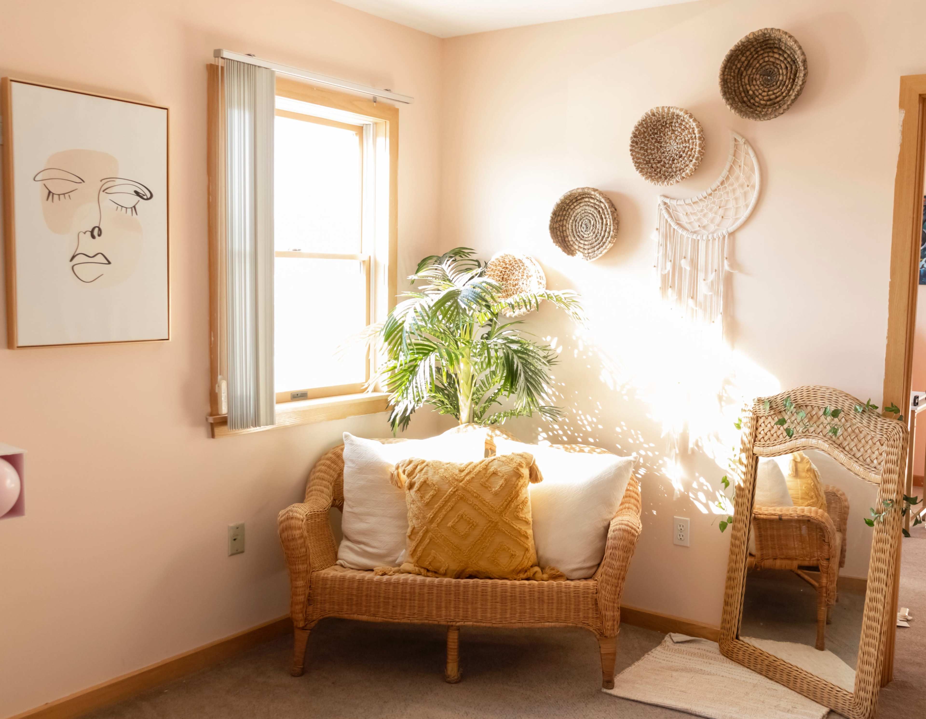 A cozy corner of a room features a rattan loveseat with decorative pillows, a potted plant, and wall hangings, illuminated by natural light streaming through a window.
