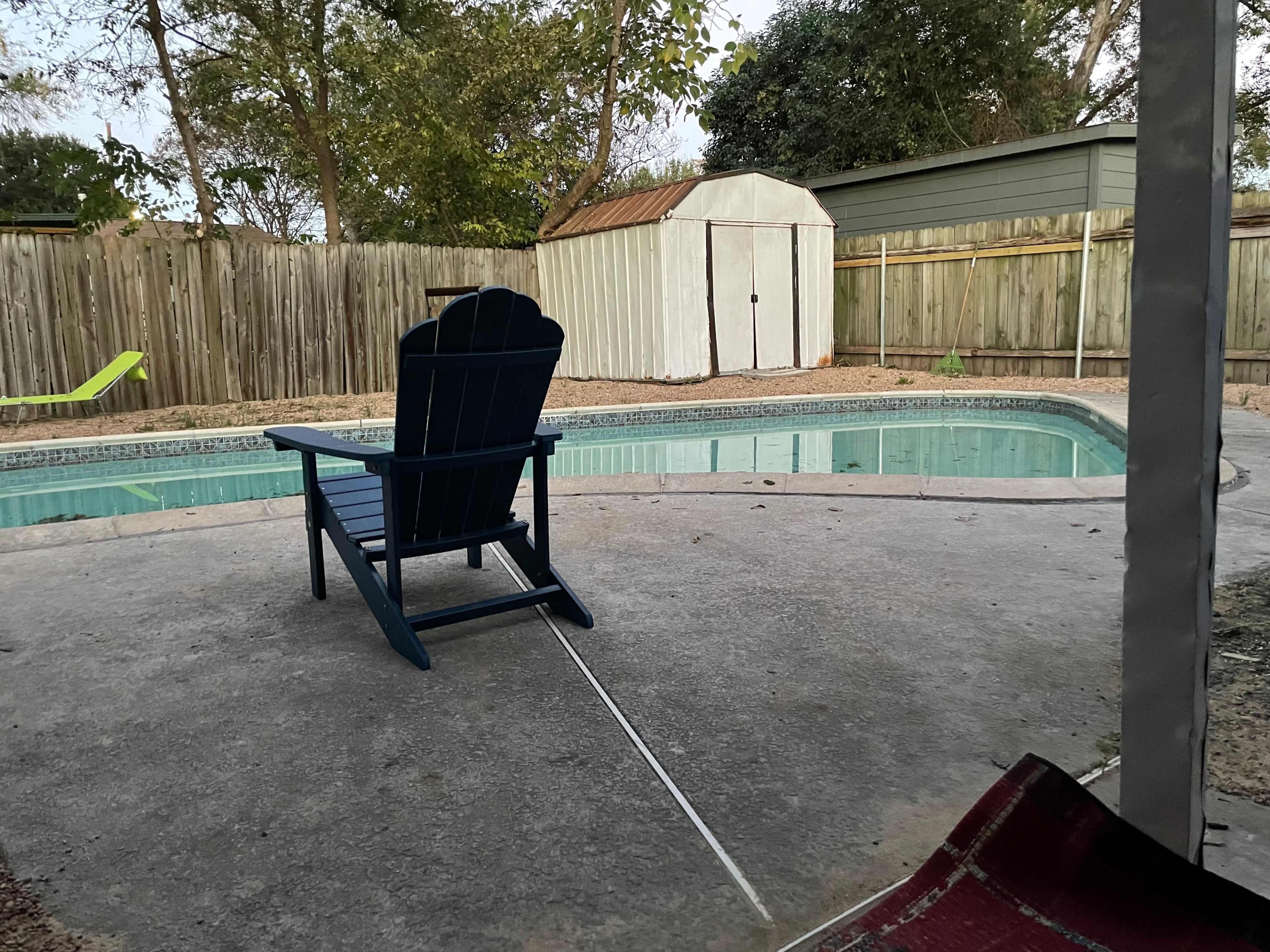 A blue Adirondack chair faces a rectangular swimming pool beside a wooden fence and a shed under a tree-lined sky.