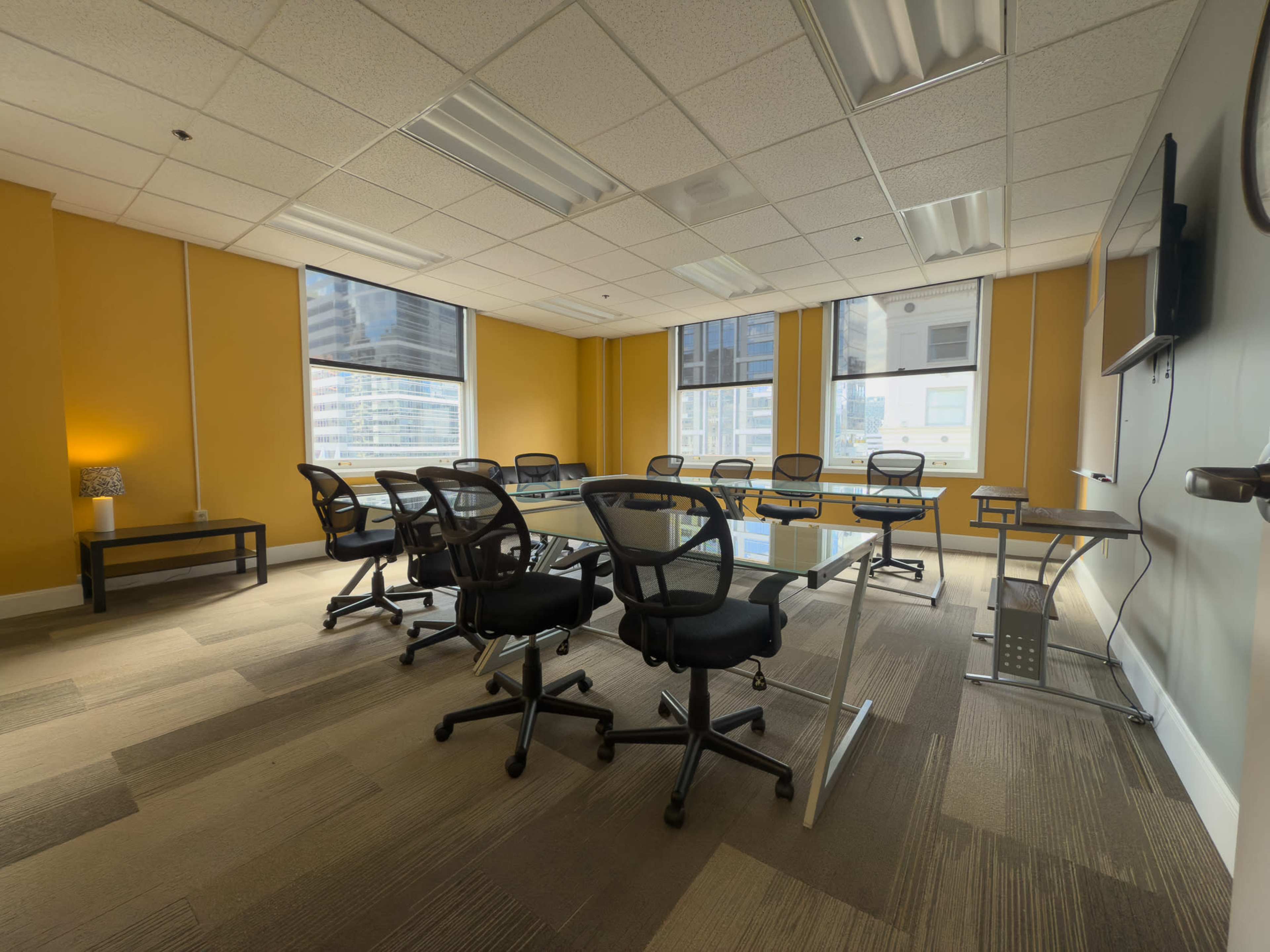 A conference room with a glass table surrounded by black office chairs, featuring yellow walls and large windows that provide natural light.