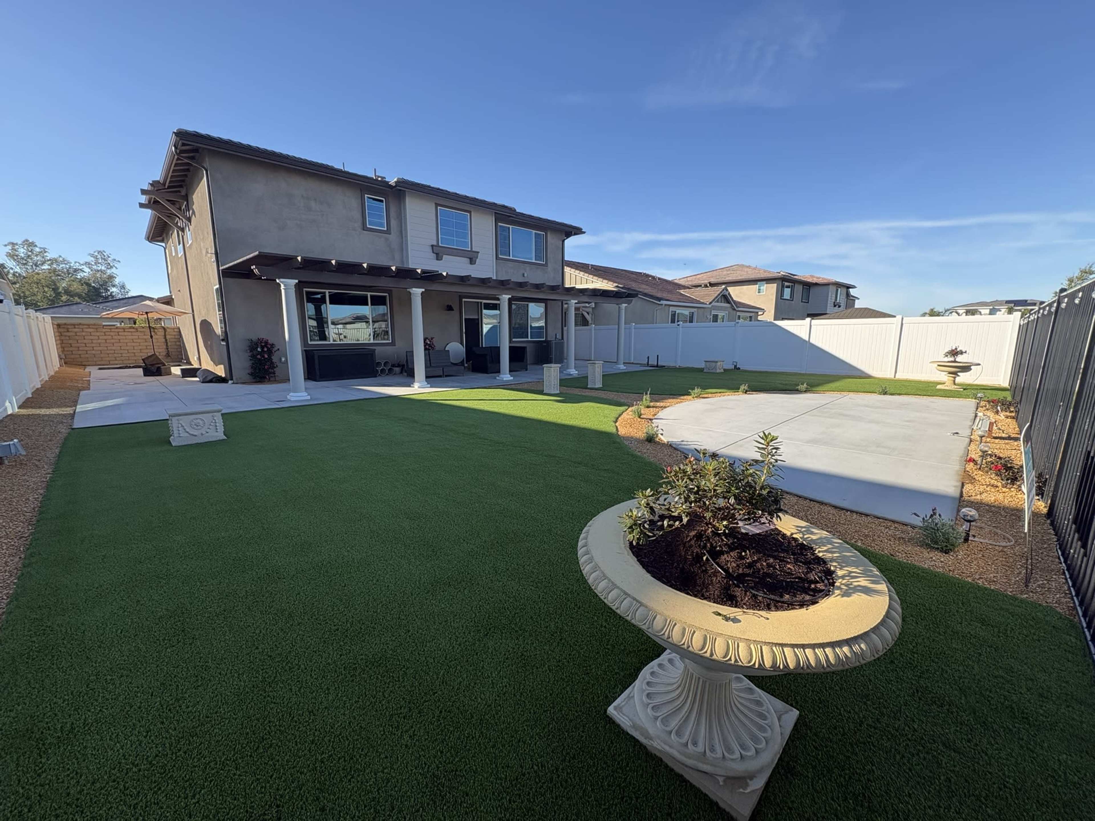 A suburban backyard features a green lawn, a concrete patio area, and a decorative stone planter in front of a two-story house.