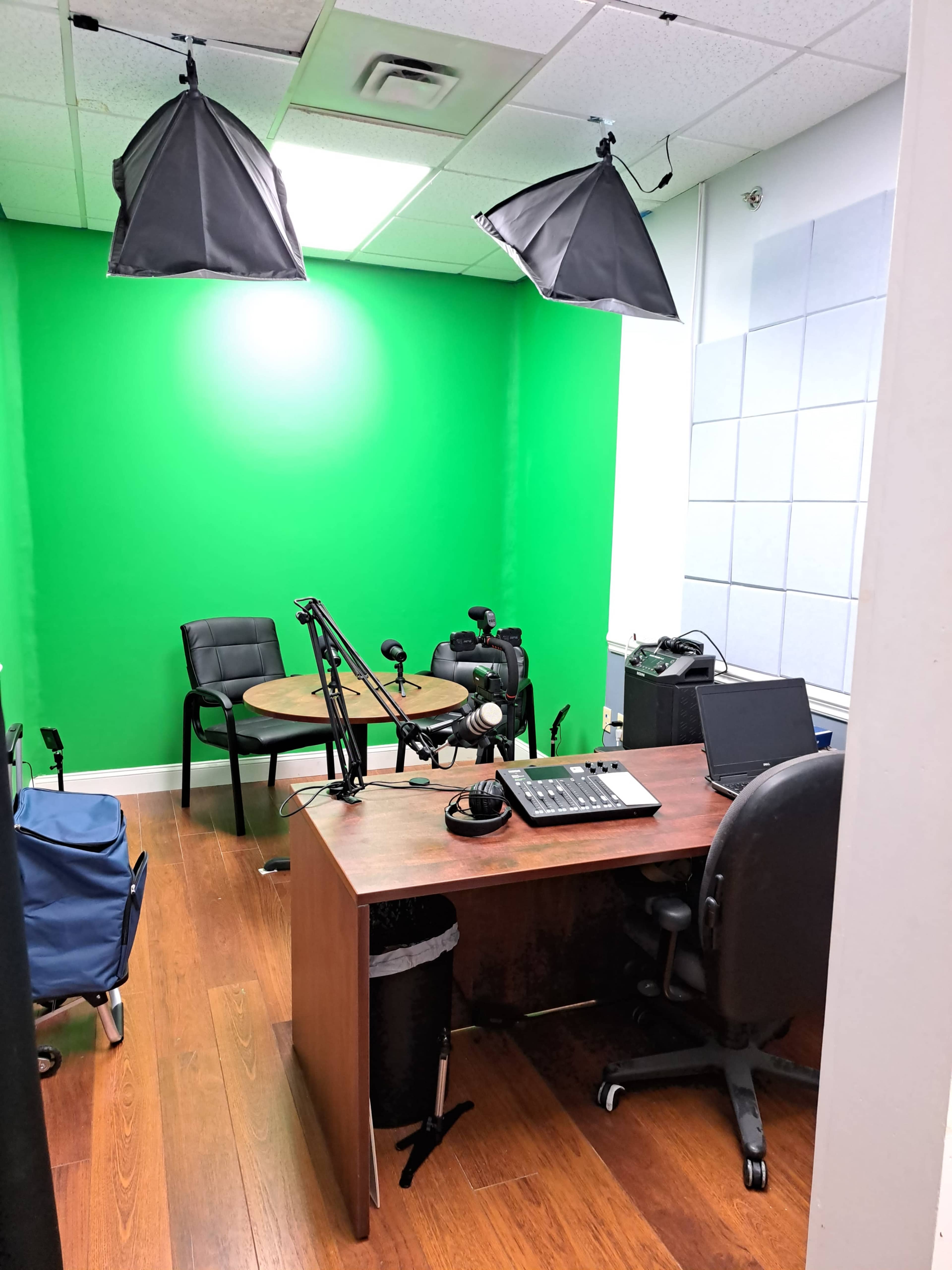 A recording studio with a green backdrop, a round table, chairs, microphones, and various recording equipment.
