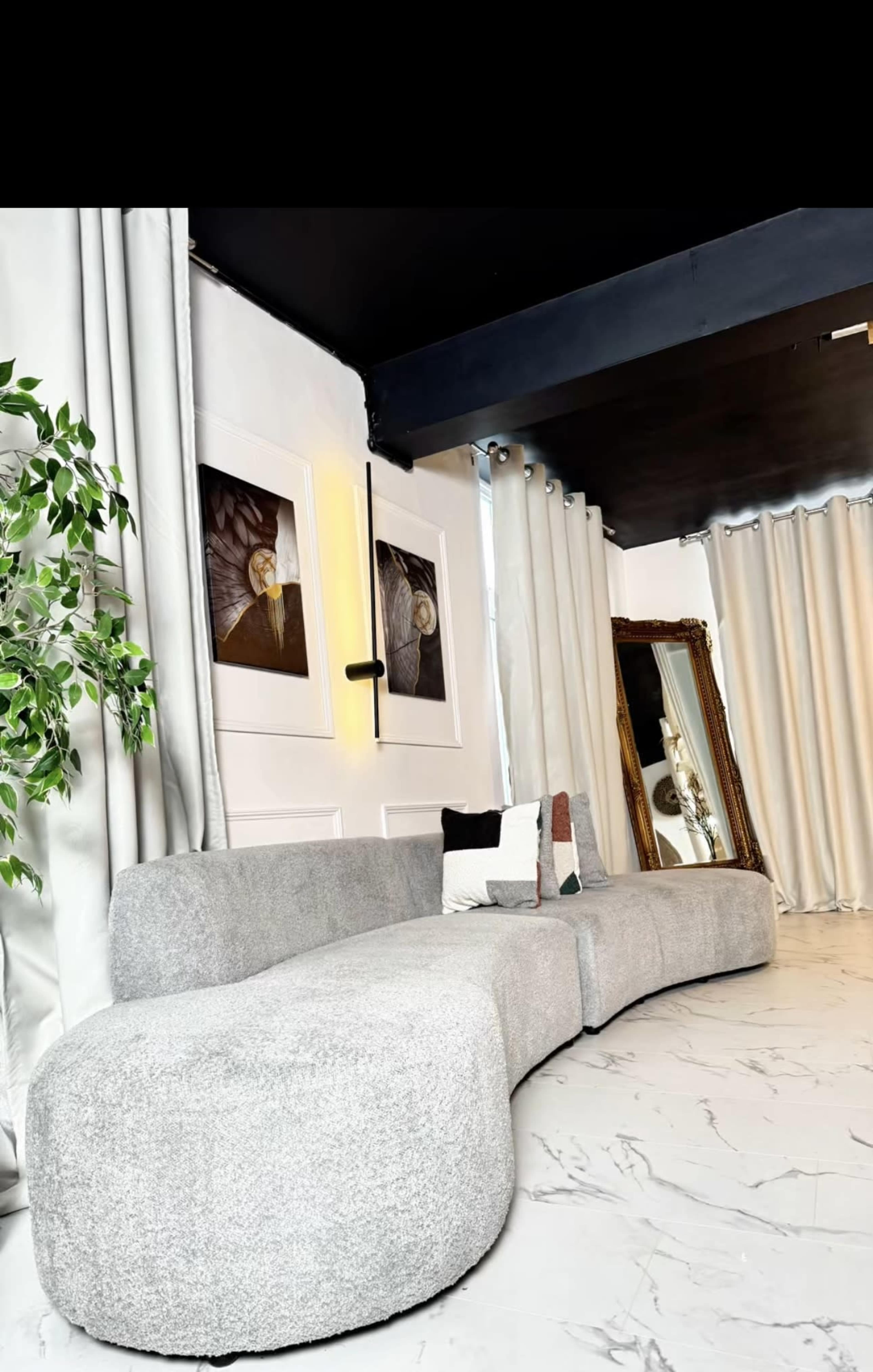 The image shows a modern living room featuring a curved gray sofa, two framed artworks on the wall, and a large mirror beside floor-to-ceiling curtains.