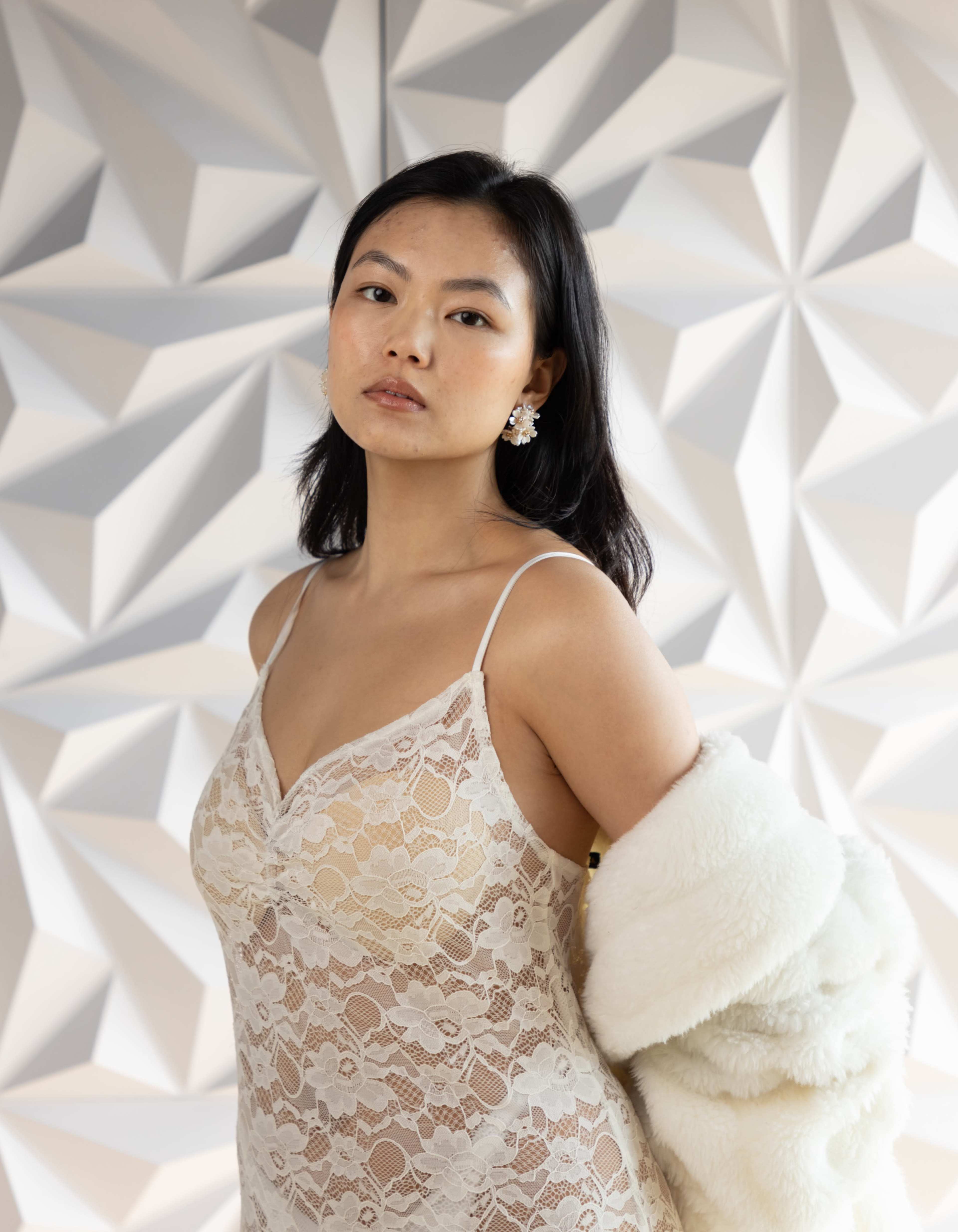 A woman poses elegantly in a lace dress while holding a fluffy white garment over her shoulder against a textured backdrop.