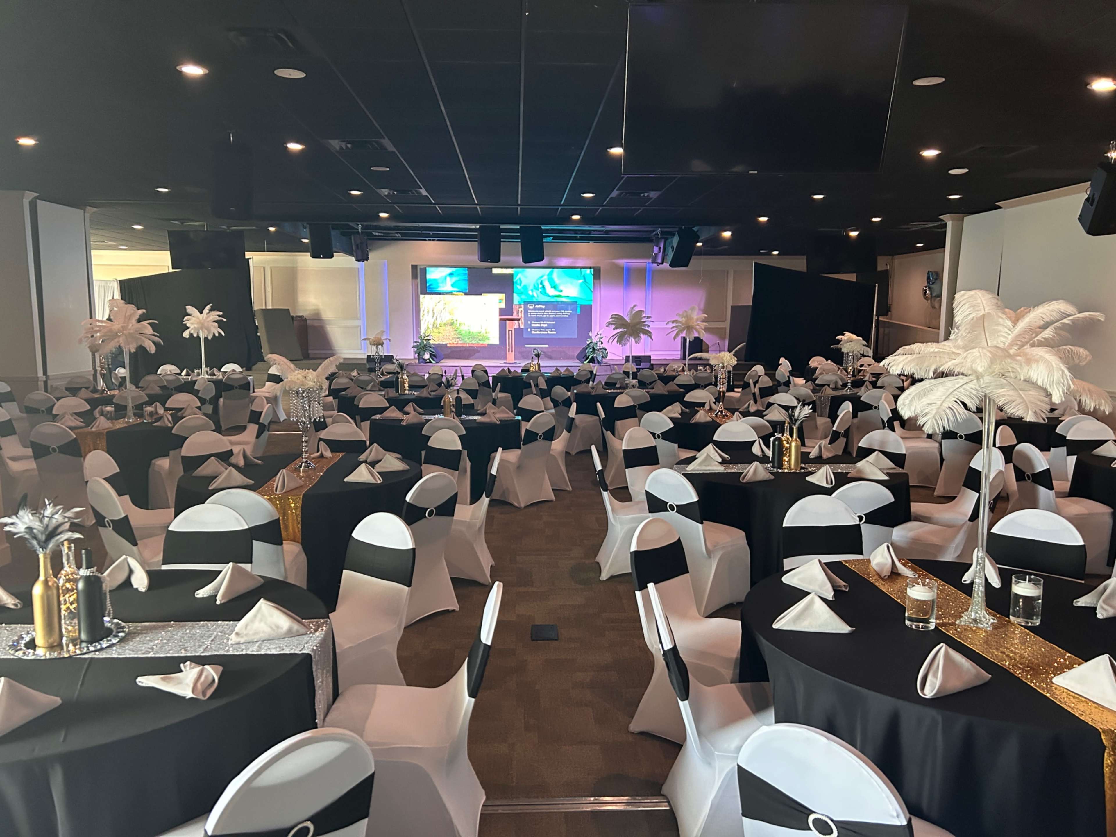 A banquet hall set for an event, featuring round tables covered in black and white tablecloths, adorned with decorative centerpieces and napkins.
