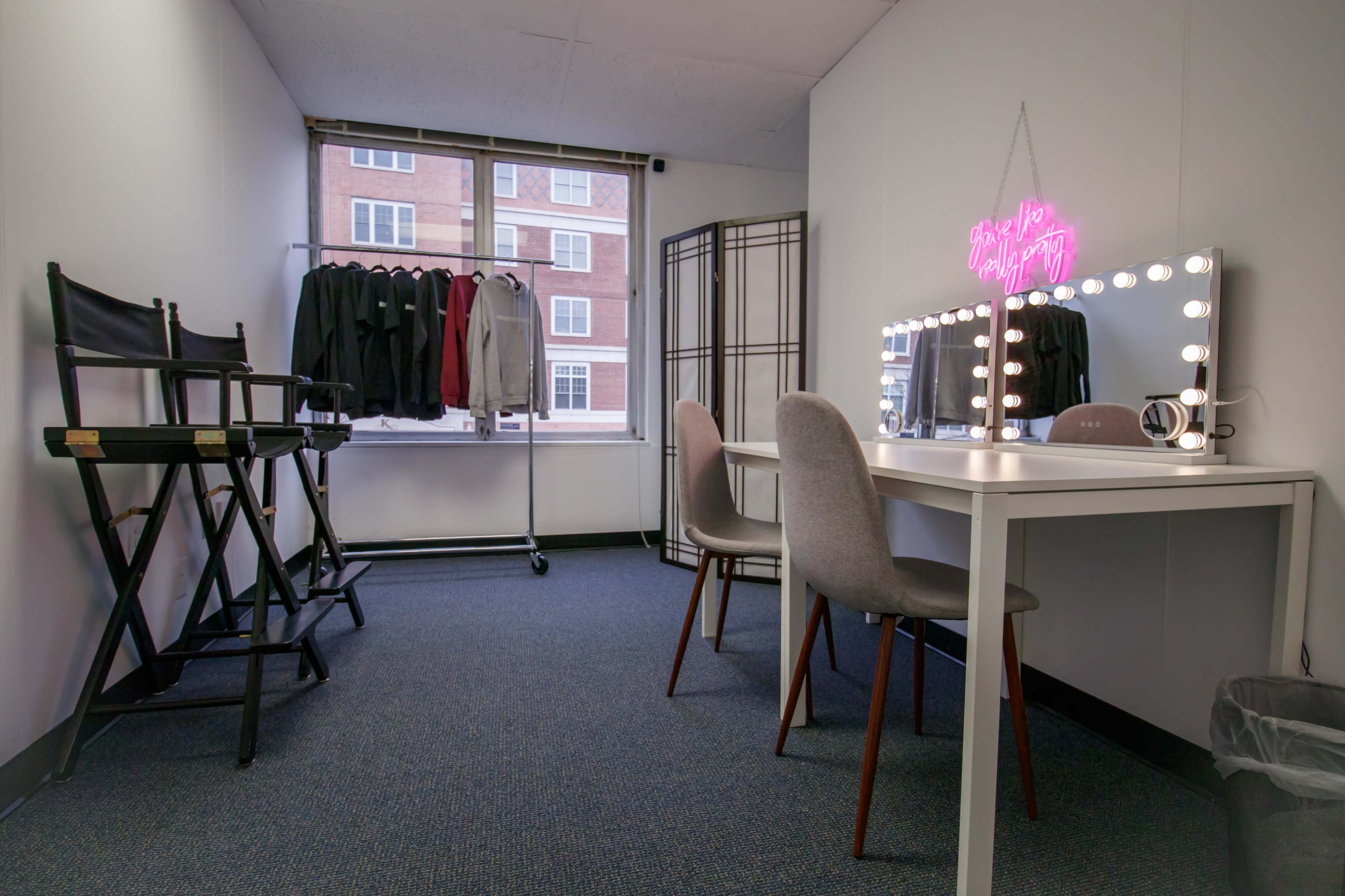 A well-lit room featuring a vanity mirror with lights, two gray chairs, a table, and a clothing rack with black garments near a window.
