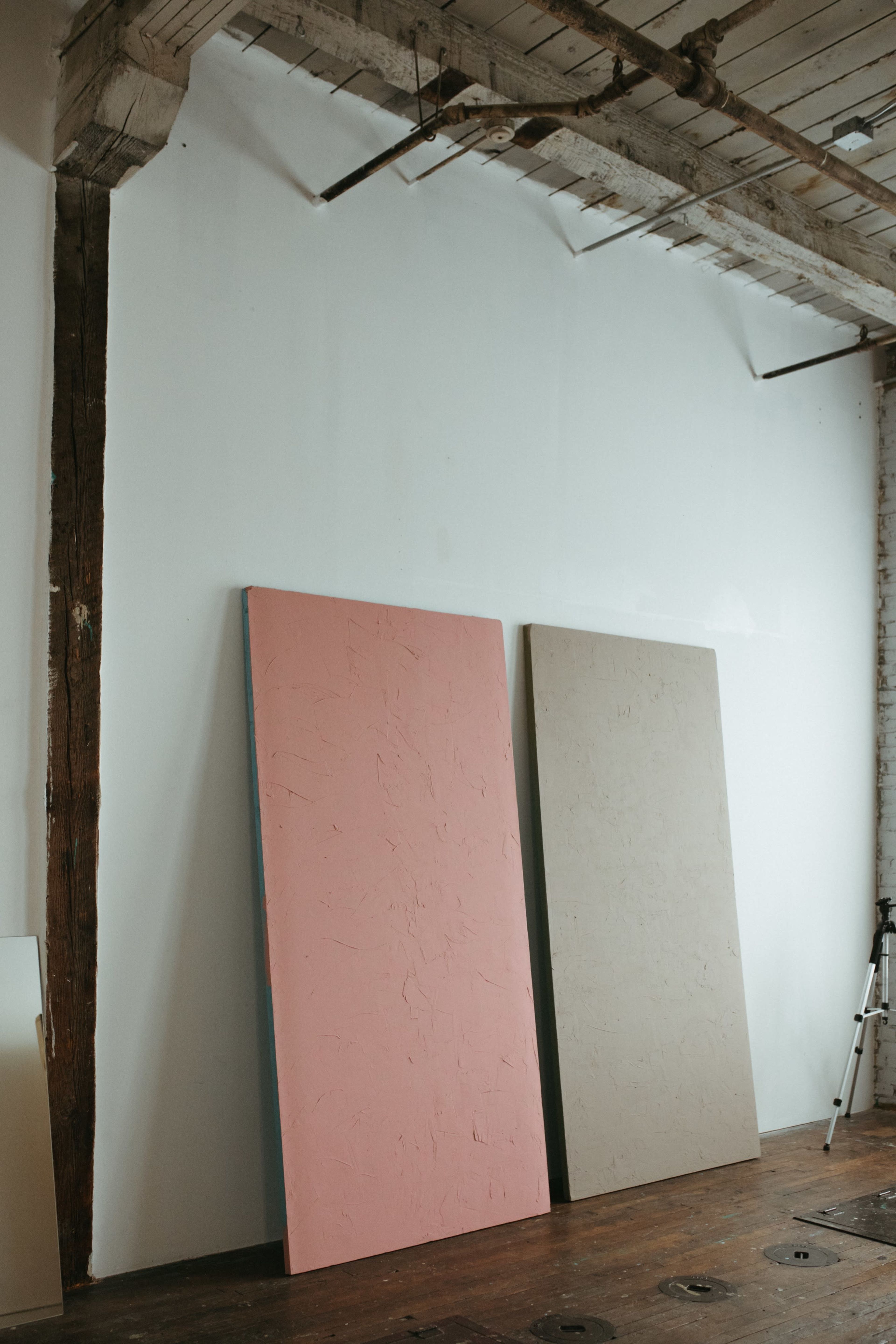Two large, unframed panels, one pink and one beige, are leaning against a white wall in a room with wooden floors and exposed beams.