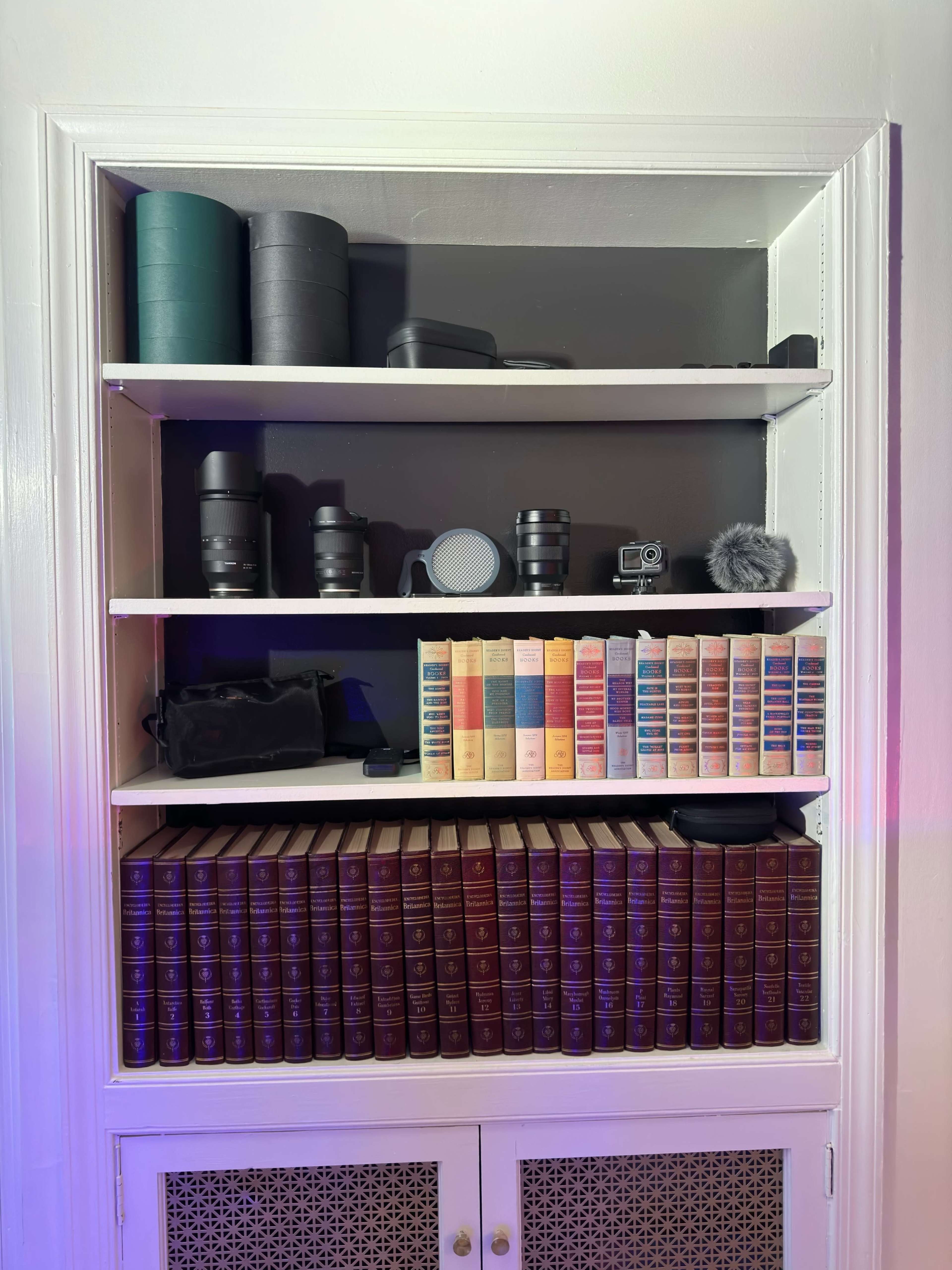 A bookshelf with photography equipment on the top two shelves and a collection of books with spines in various colors on the bottom shelf.