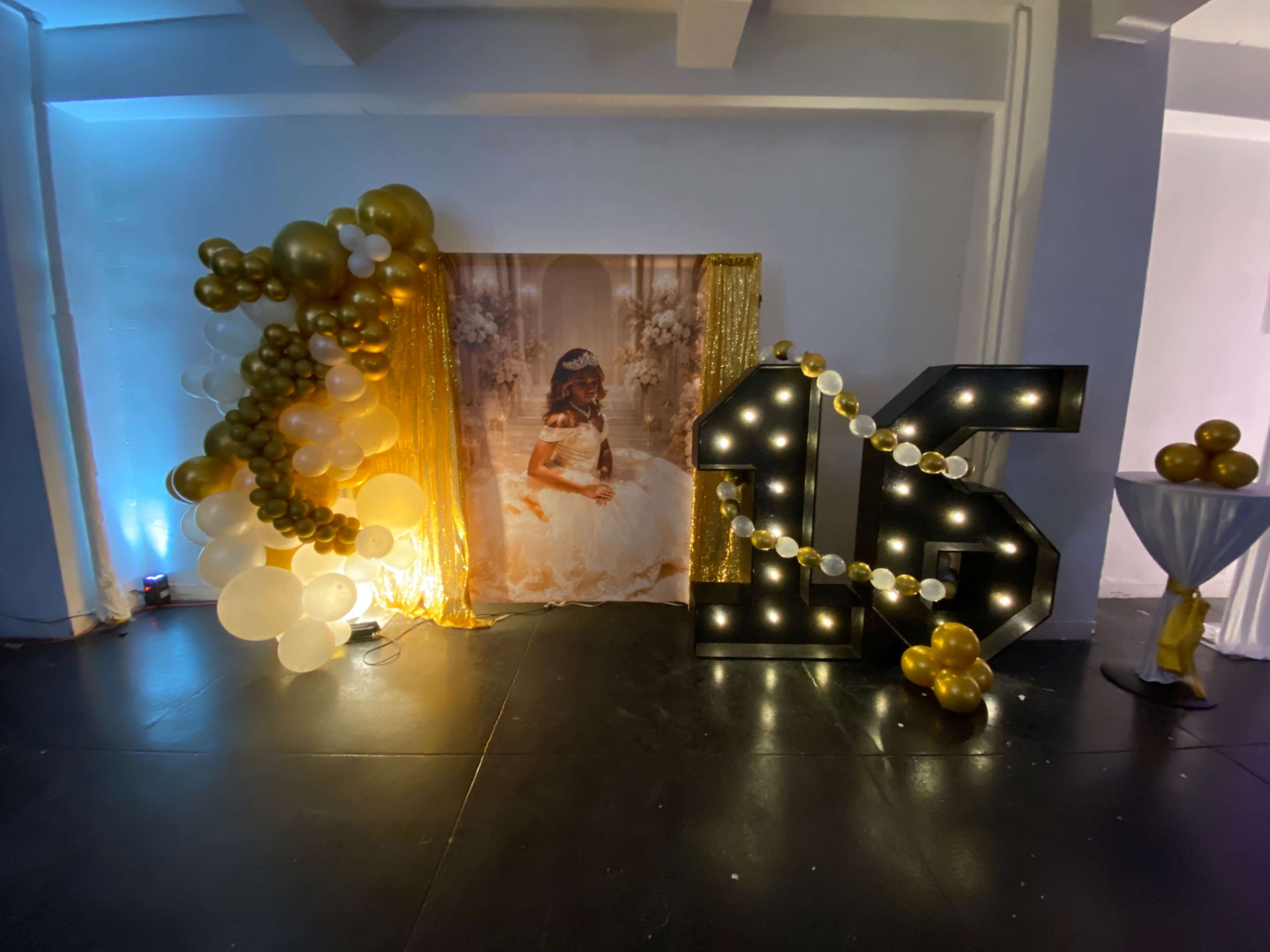 The scene features a decorated event space with a large illuminated "16" sign, a balloon arrangement, and a backdrop showcasing a portrait of a girl in a gown.