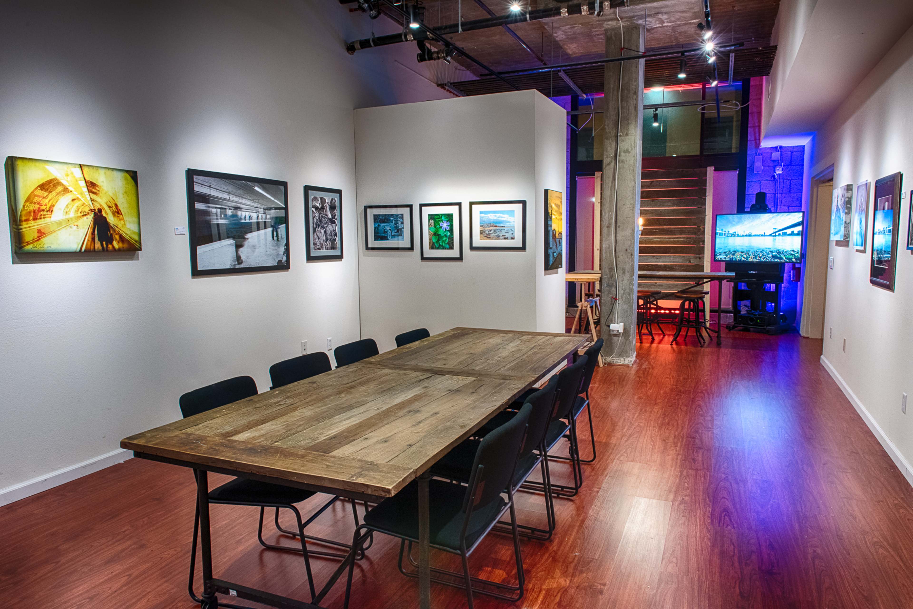 A spacious gallery features a long wooden table surrounded by black chairs, with framed photographs displayed on the walls and a staircase leading to an upper level.