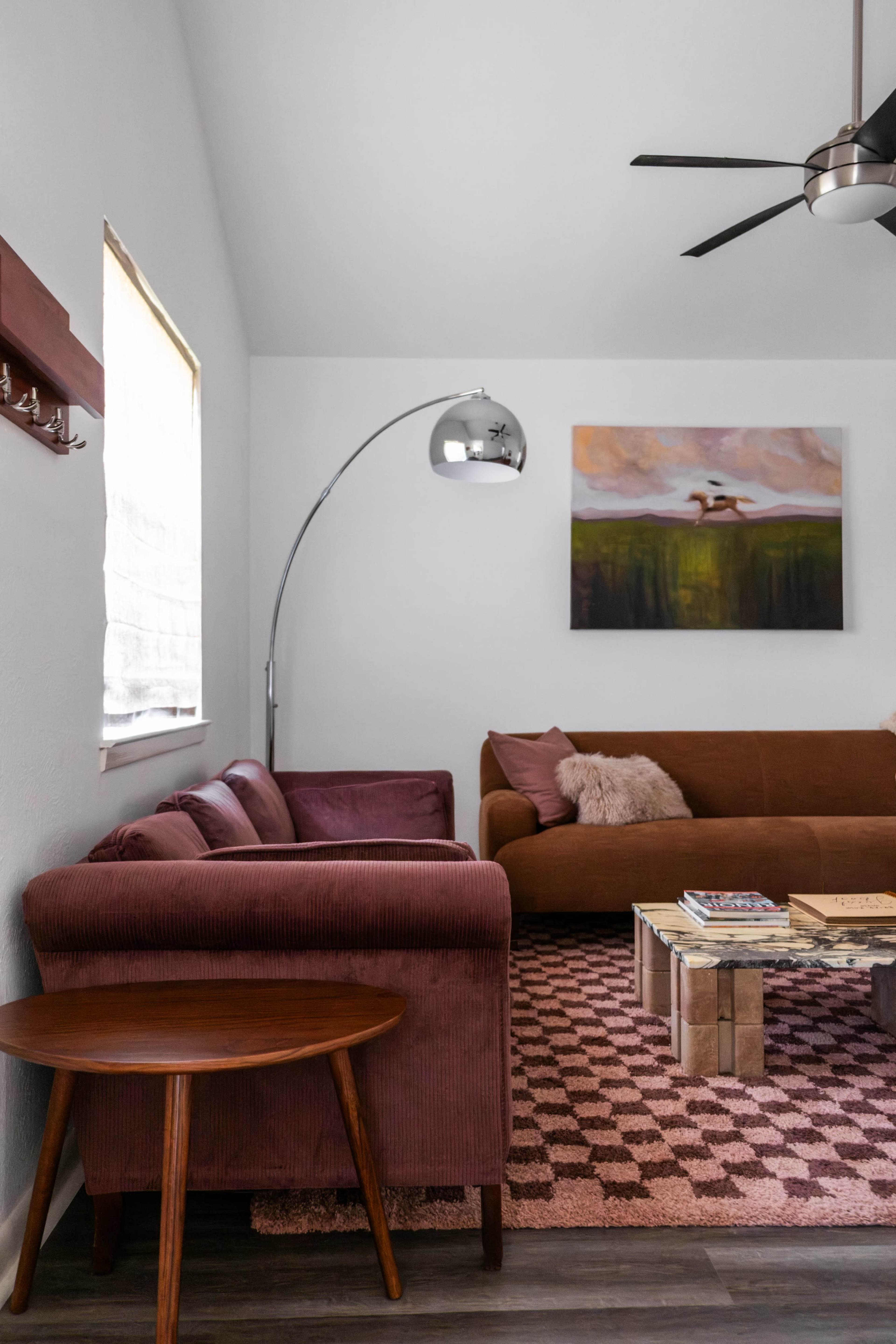 The image shows a modern living room with a brown sofa, a burgundy armchair, a circular wooden table, and a floor lamp, accented by a colorful painting on the wall.