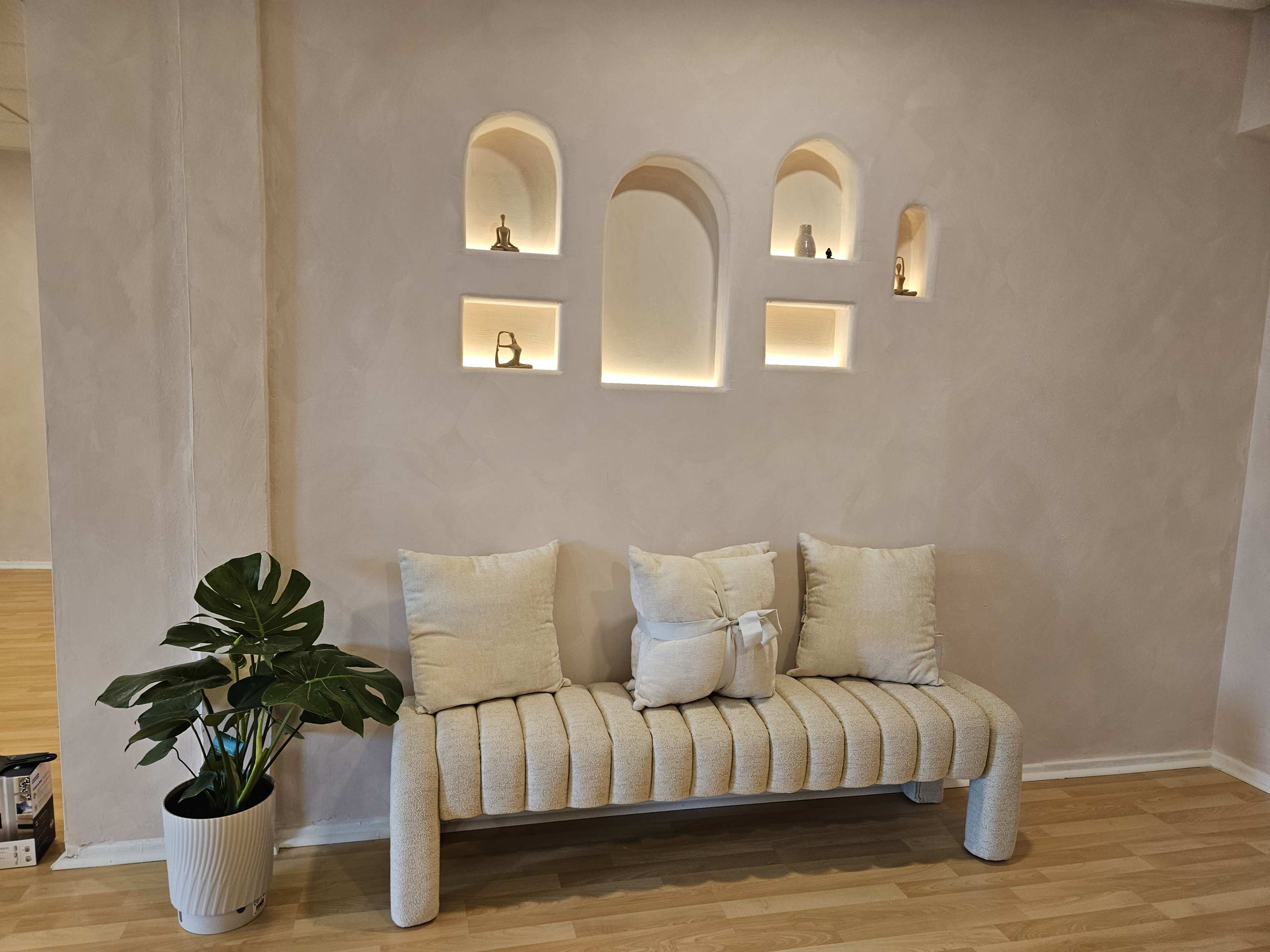 The image shows a minimalist interior with a light-colored wall featuring arched niches that are backlit, along with a textured sofa and a potted plant.