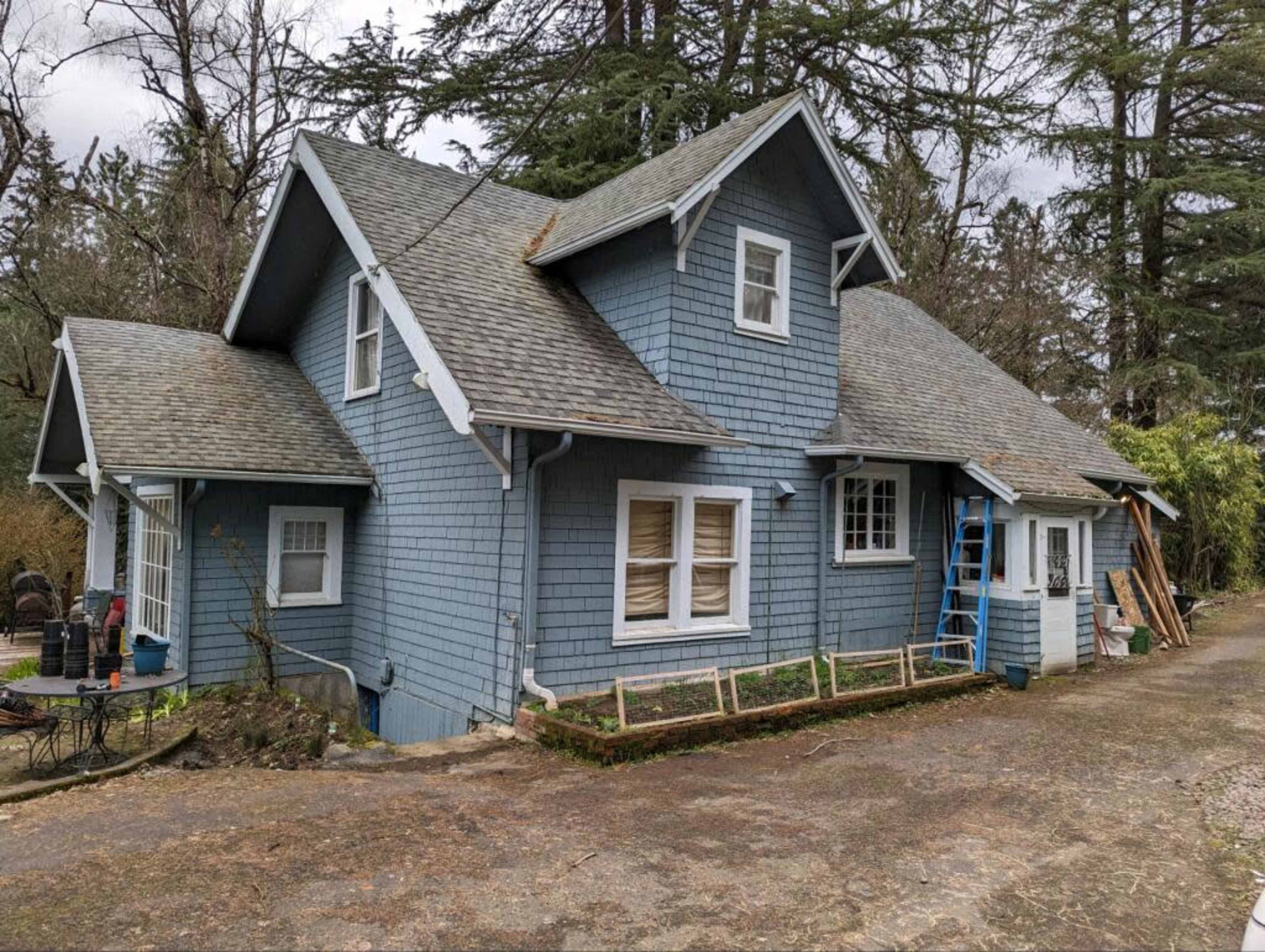 A two-story blue house with a gabled roof and a side porch is situated amidst trees, with ladders and construction materials nearby.