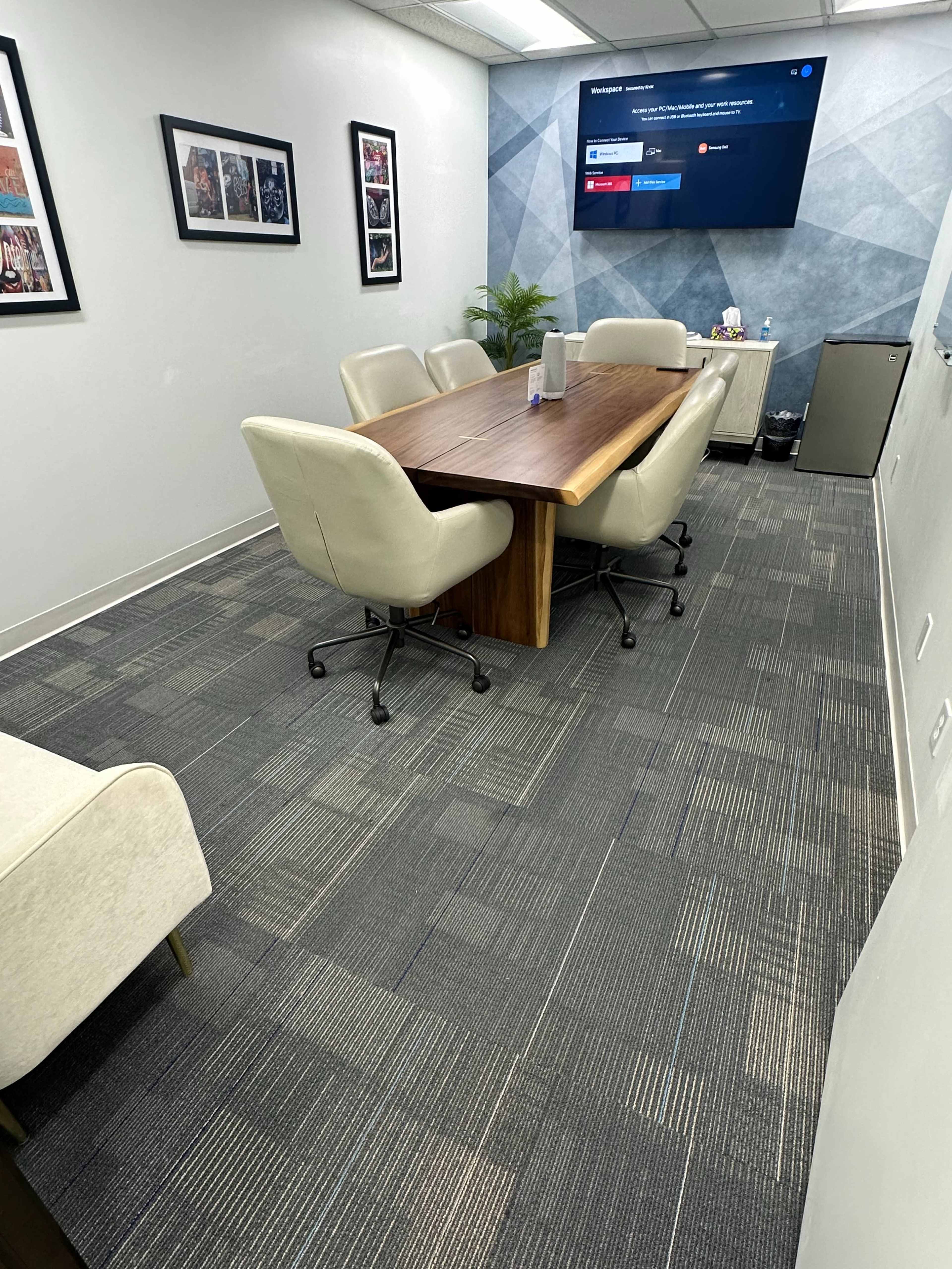A conference room features a large wooden table surrounded by four upholstered chairs, with a TV mounted on the wall displaying a screen.