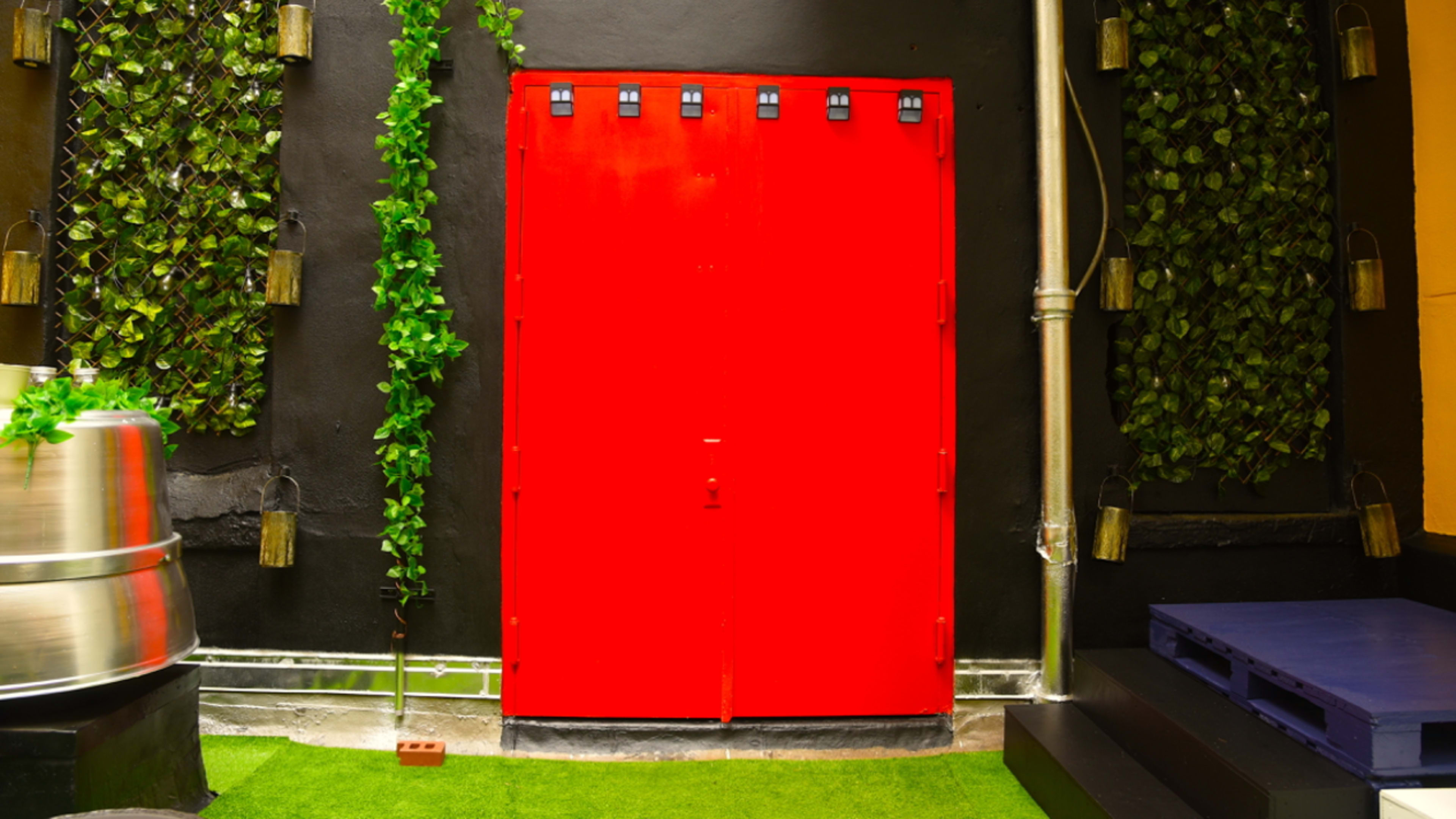 A bright red door is framed by green foliage on a black wall, with metal accents and a silver cylindrical structure nearby.