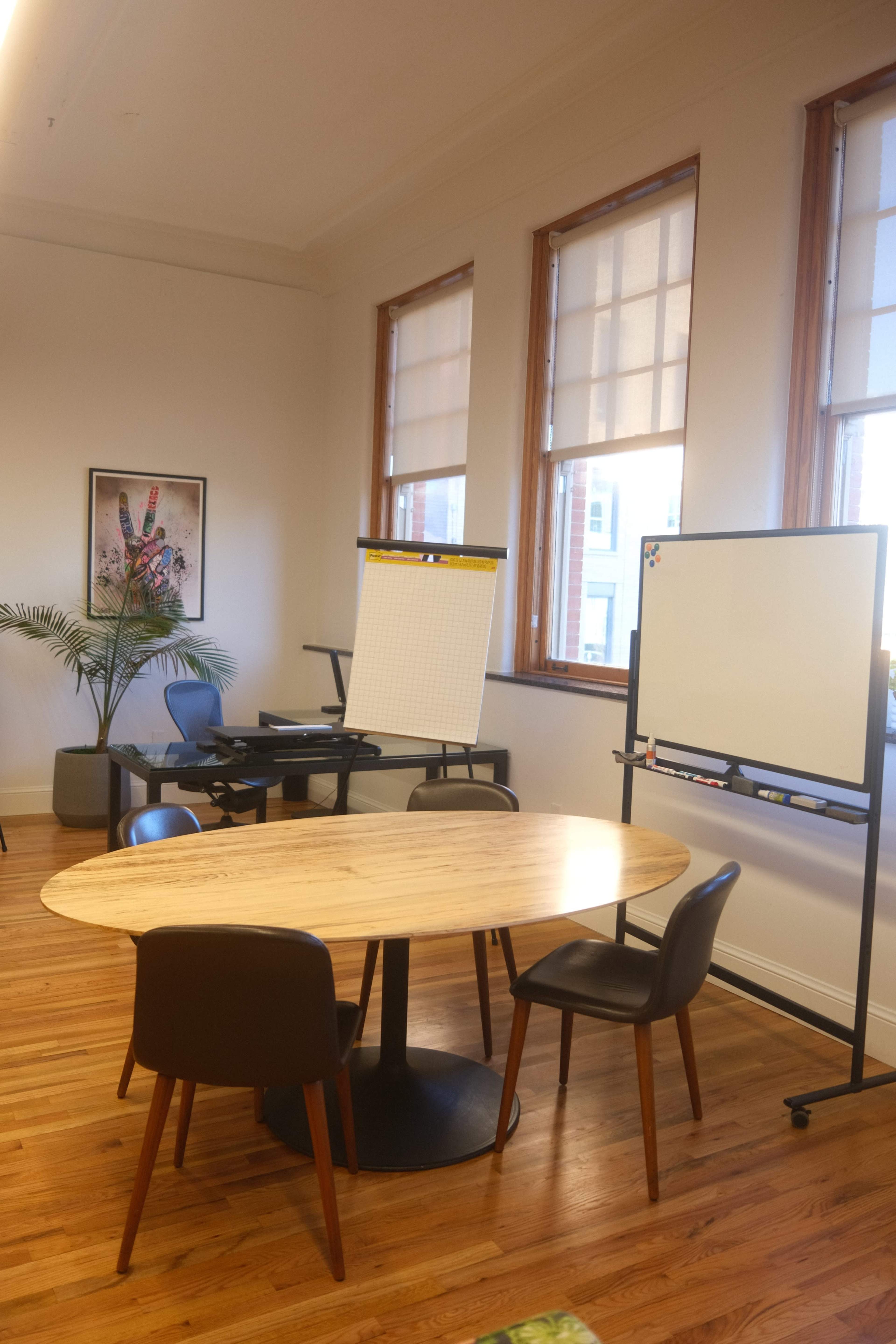 The image shows a small meeting room with a round wooden table, several chairs, and whiteboards positioned against the walls.