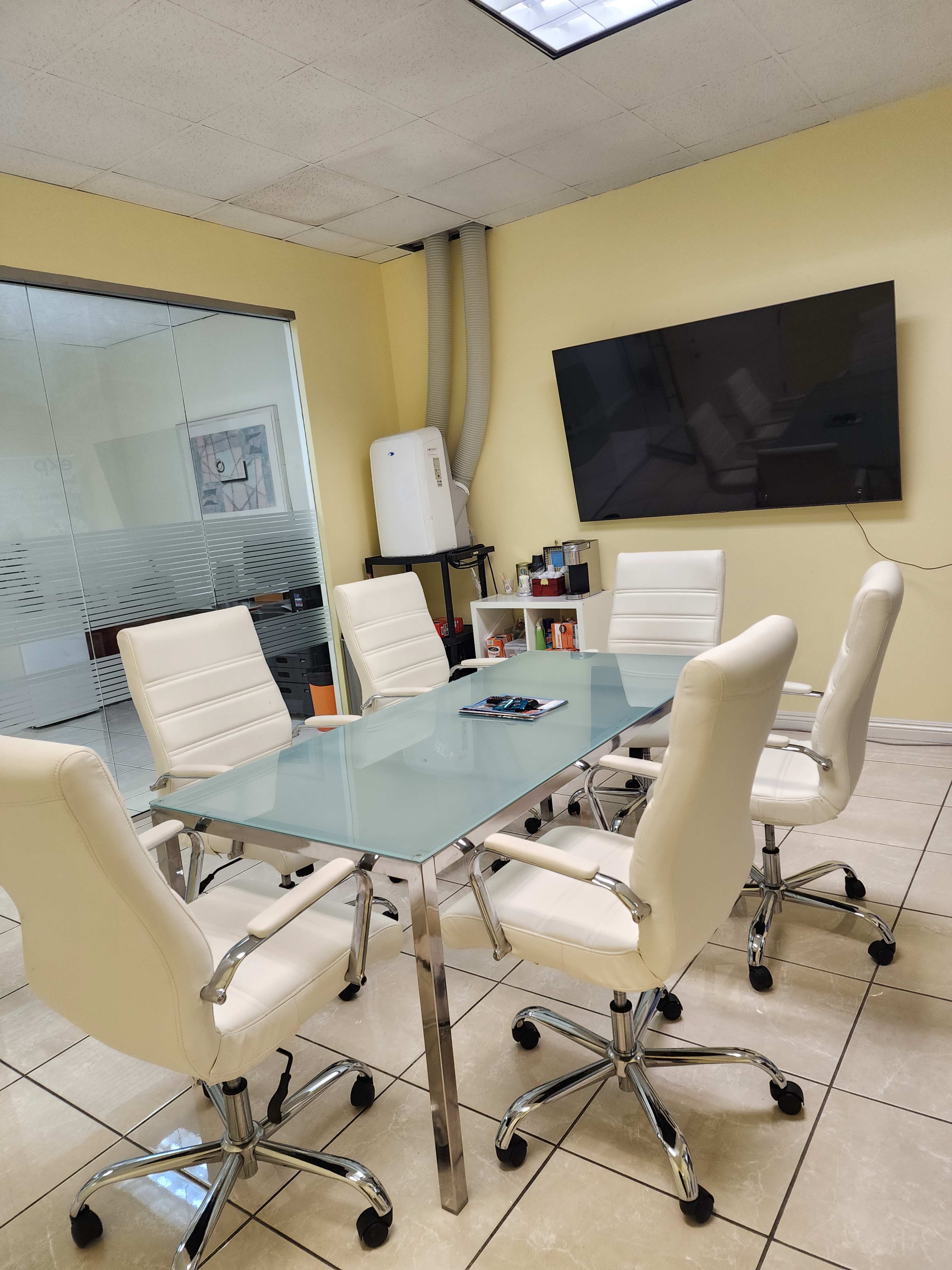 The image shows a meeting room with a glass table surrounded by six white chairs, a wall-mounted television, and a window with frosted glass panels.