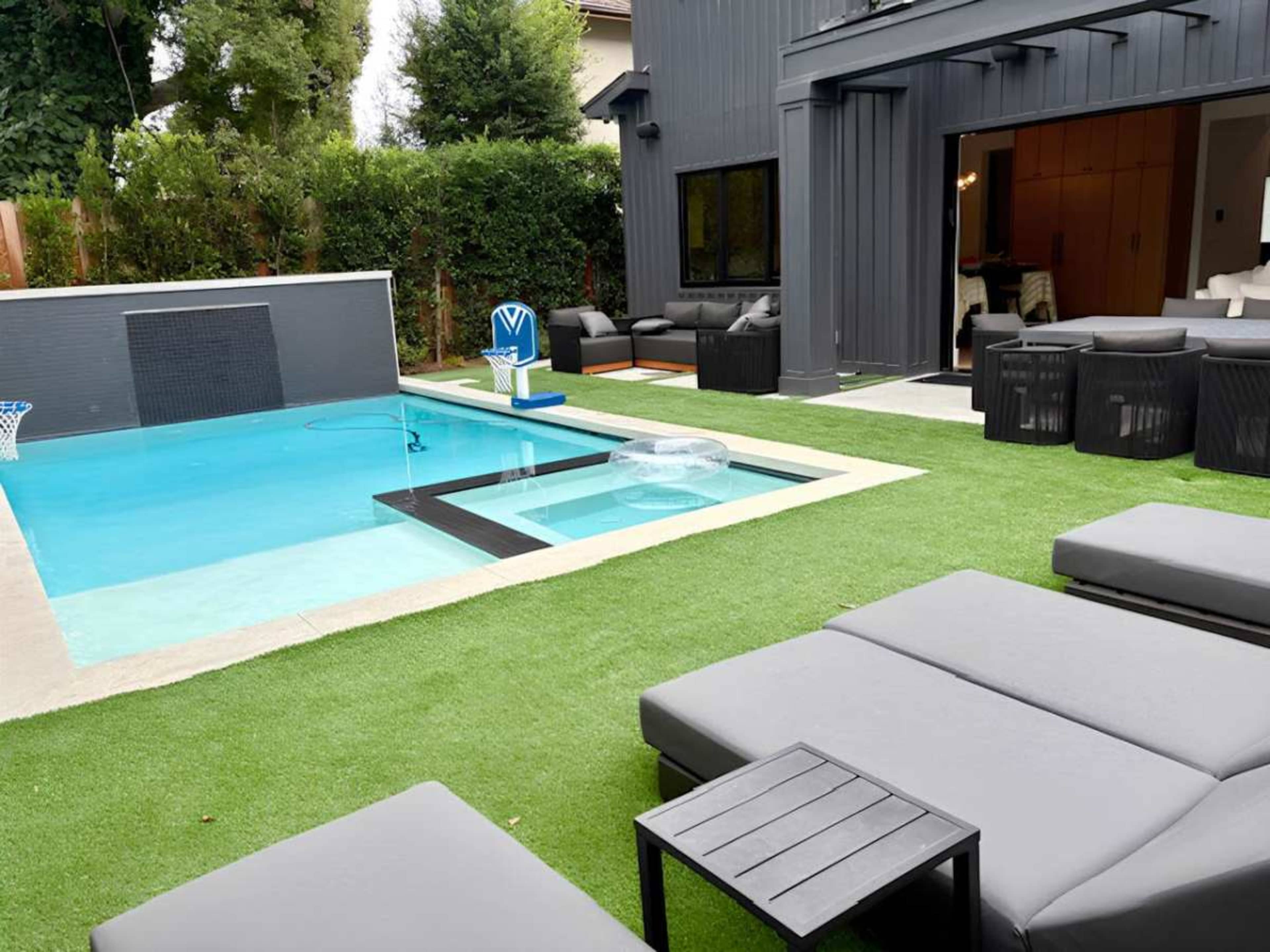 The image shows a backyard with a modern swimming pool, lounge chairs, and a basketball hoop near a well-manicured lawn.