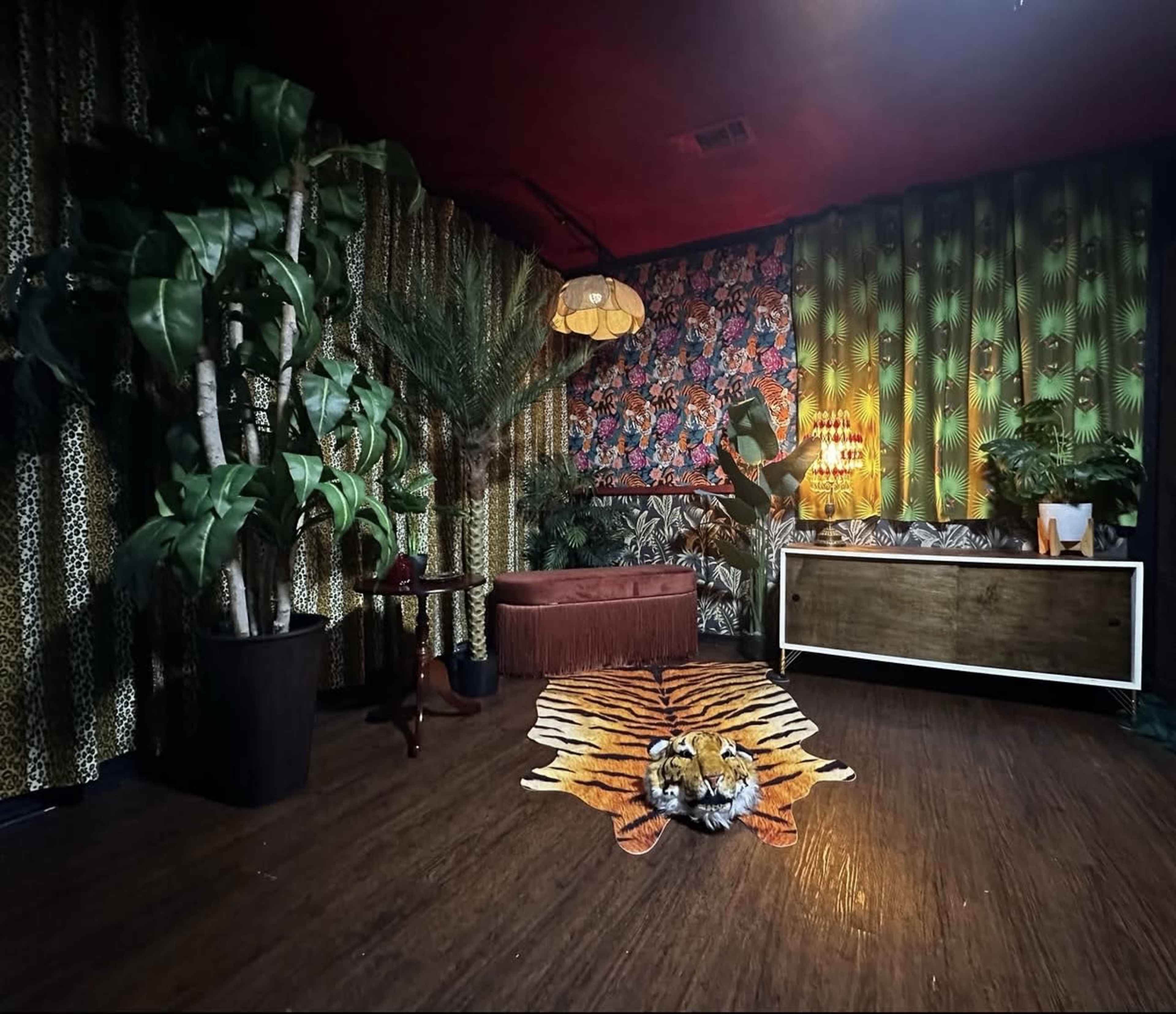 The scene features a stylish room with patterned wallpaper, a tiger-striped rug, indoor plants, a vintage armchair, and an ornate light fixture.