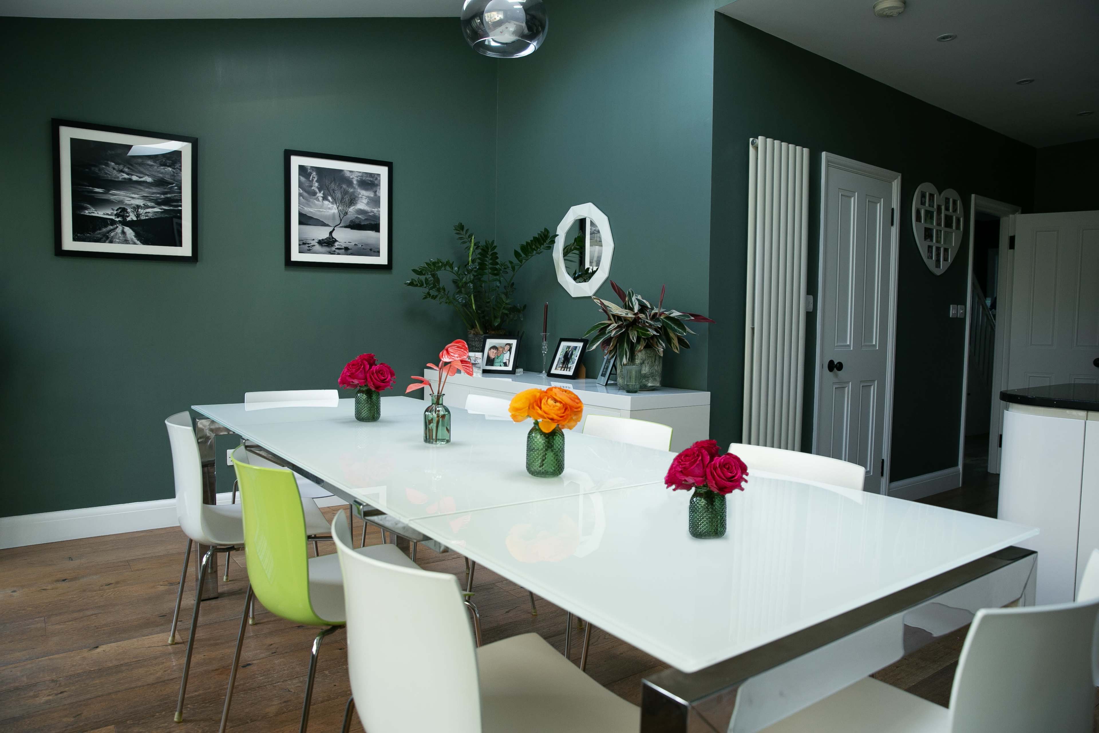 A modern dining room features a long white table set with vases of flowers, surrounded by a mix of white and green chairs, with green walls and framed photos on display.