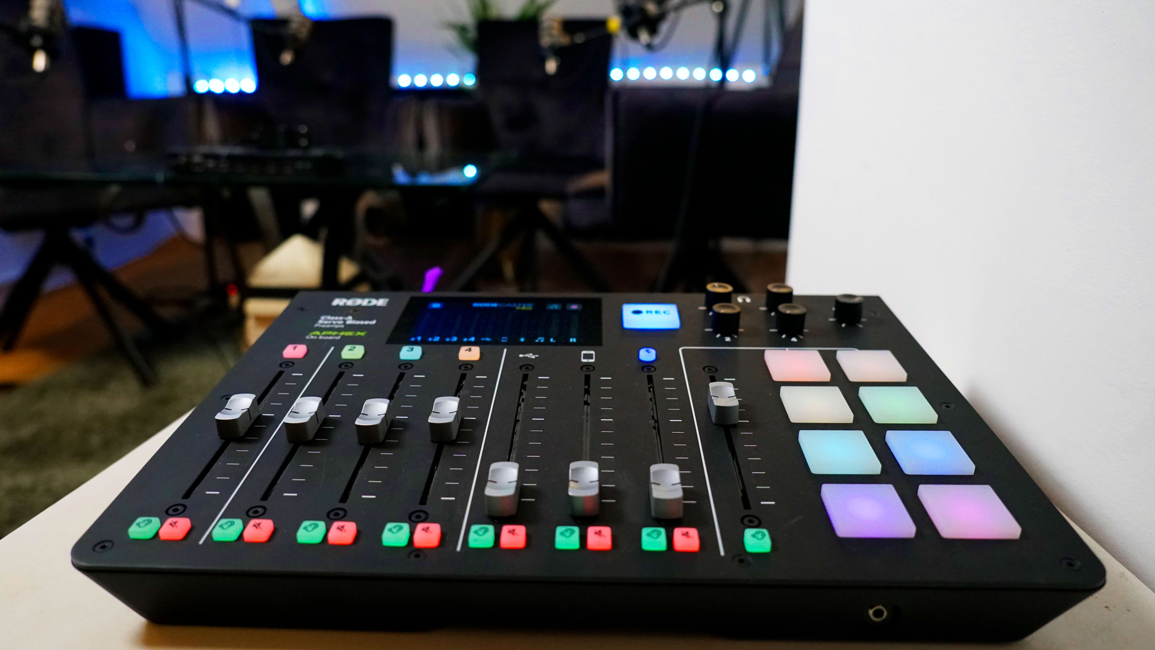A sound mixing console with faders, buttons, and pads is positioned on a table in a room with chairs and ambient lighting in the background.