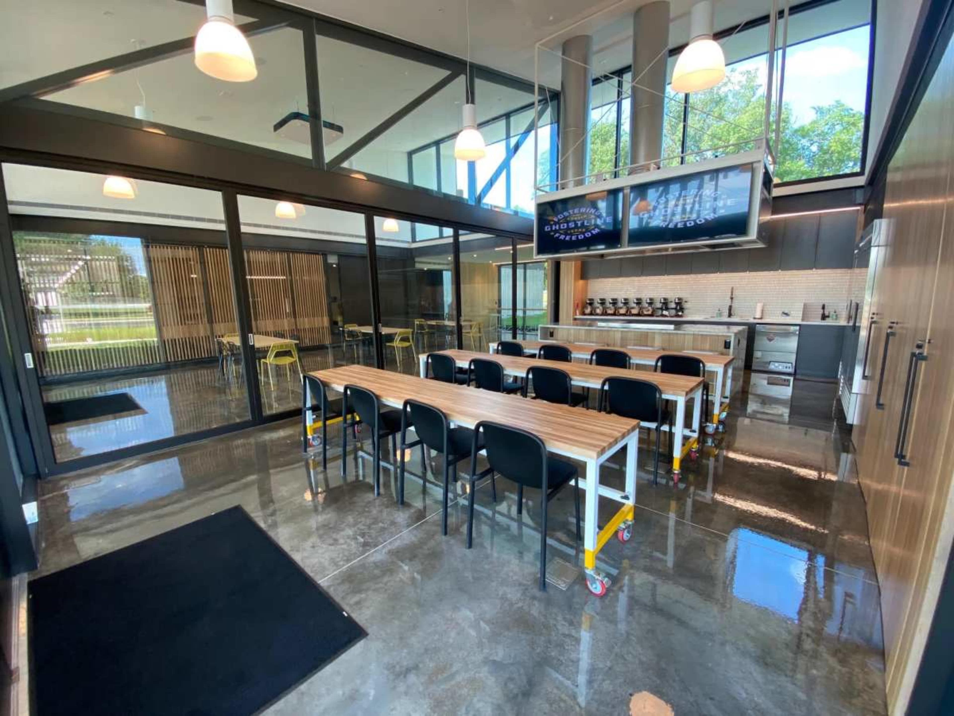 The image shows a modern kitchen and dining area with a long wooden table, black chairs, and a large television mounted on the wall.