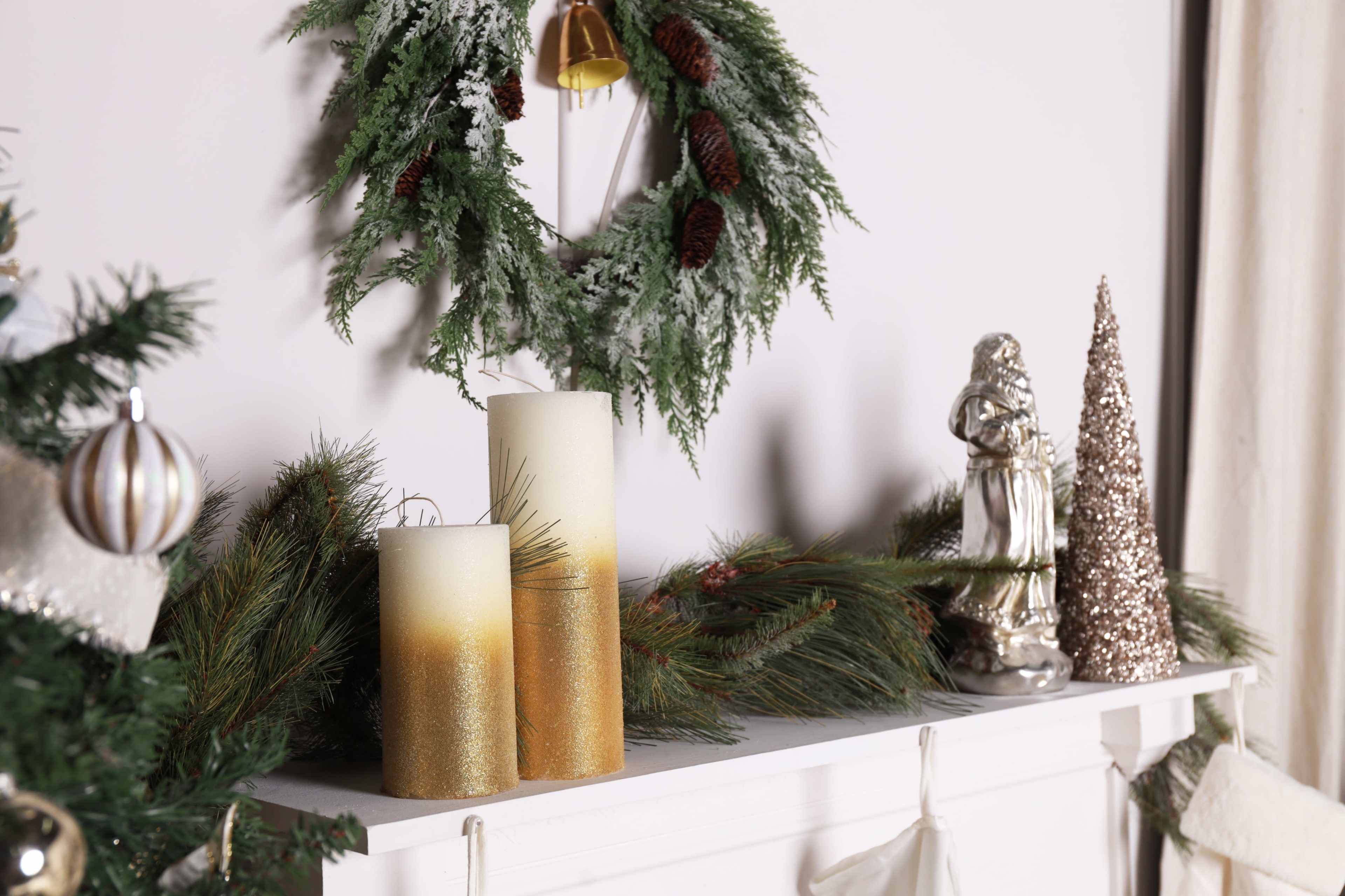 The image shows a decorated mantel featuring two gold and white candles, a green wreath with pinecones, a silver statue, and festive greenery.