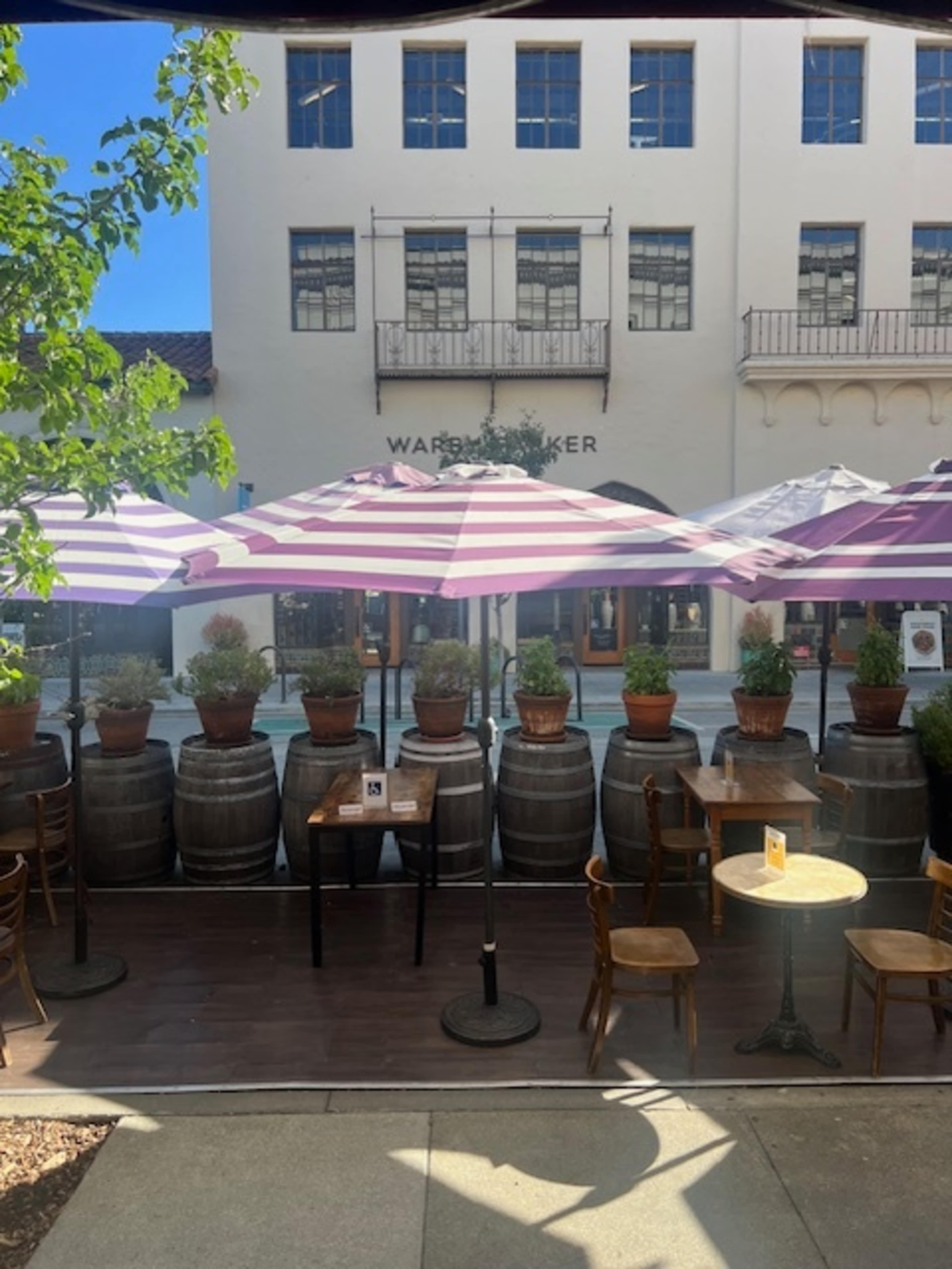 A row of wooden tables and chairs with pink and white striped umbrellas is set outside a building with large windows and a sign that reads "Warden Baker."