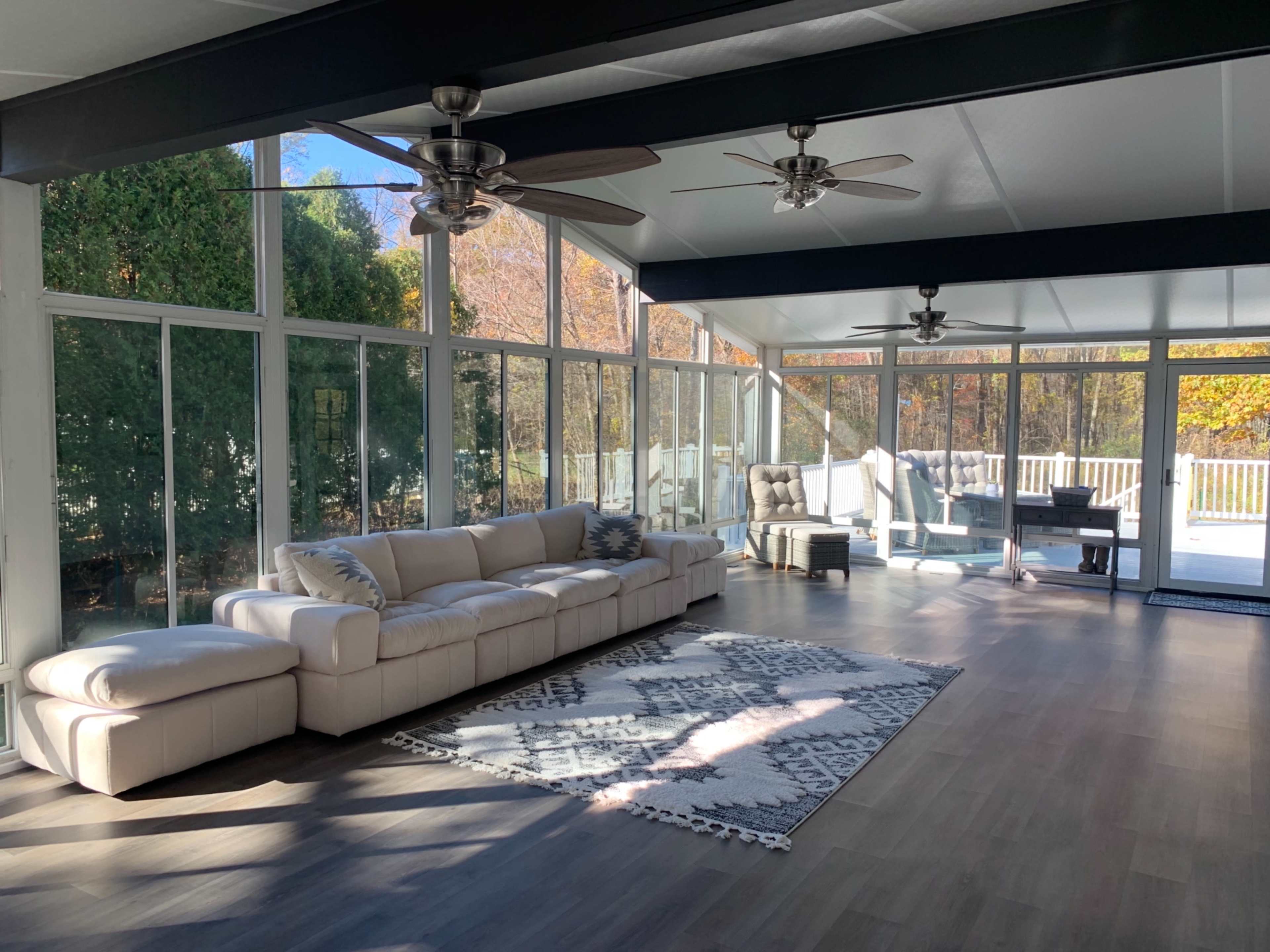 A spacious sunroom with large glass windows, two ceiling fans, a light-colored sectional sofa, and a patterned area rug on the wooden floor.